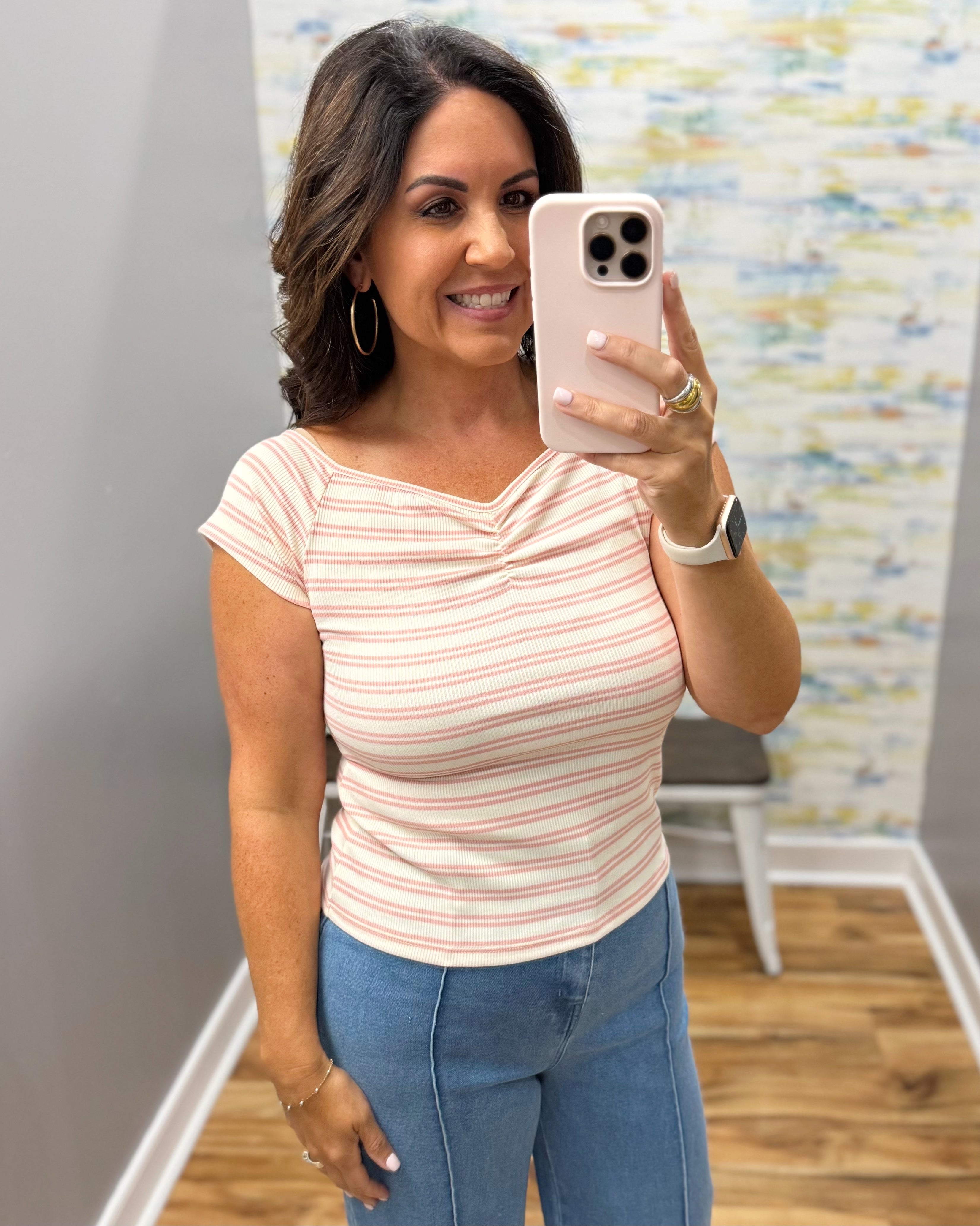 A woman taking a mirror selfie in a fitting room wearing a light pink and white striped short-sleeve top with a ruched V-neckline, paired with blue jeans.