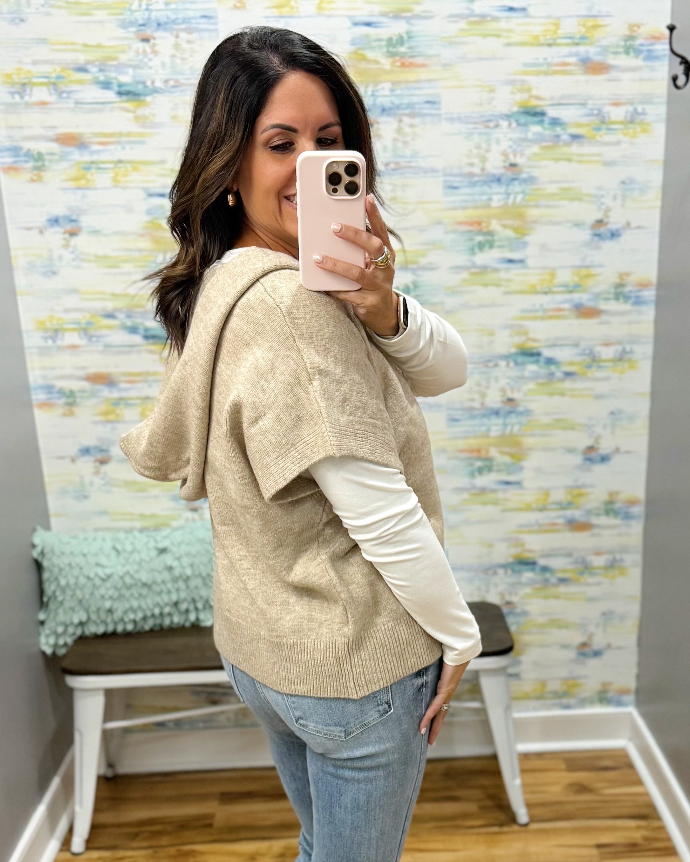 A woman takes a mirror selfie wearing a beige knit hooded poncho layered over a white long-sleeve top and light blue jeans, standing in front of a patterned wallpaper background.