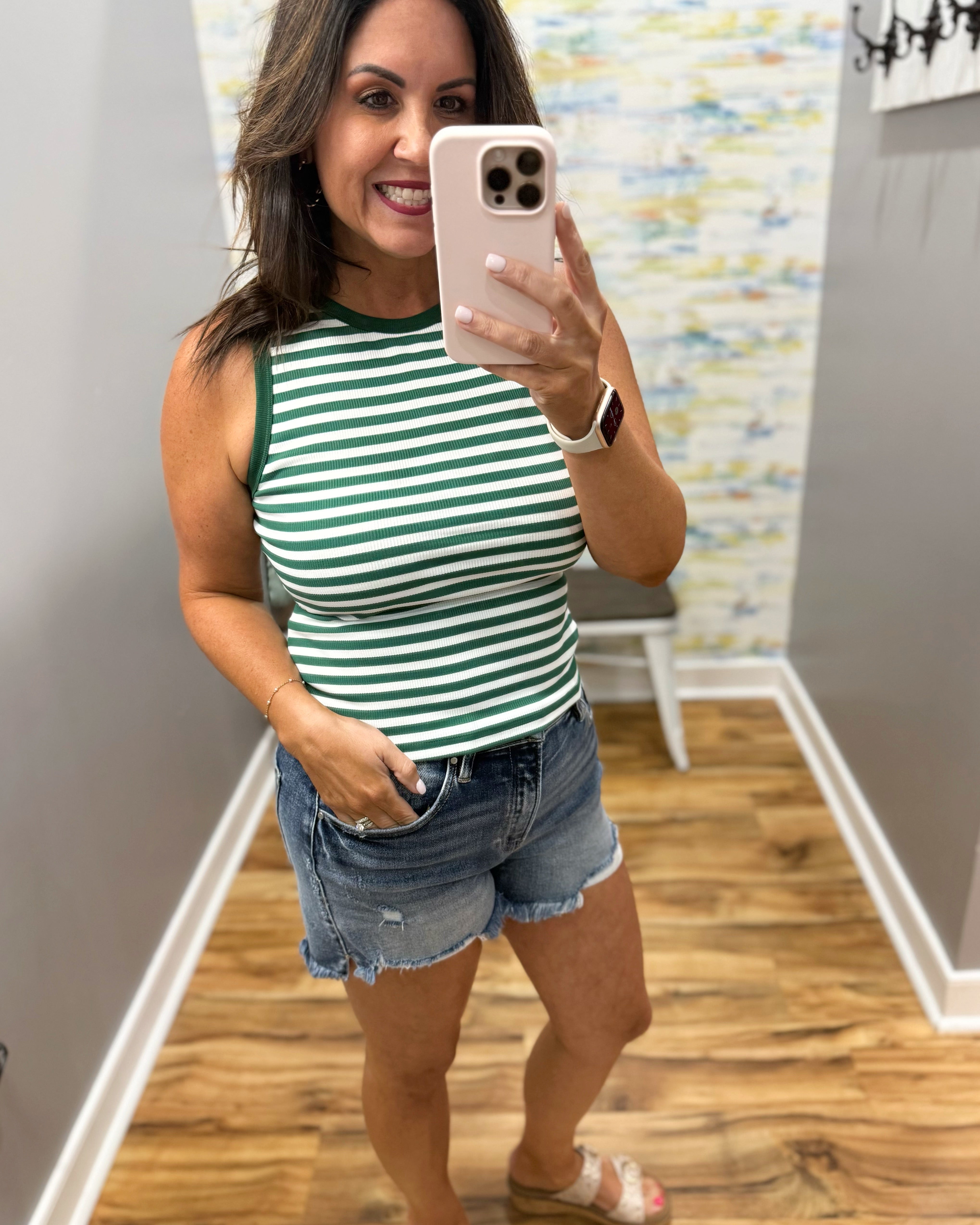 Woman taking a mirror selfie wearing a green and white striped tank top and denim shorts.