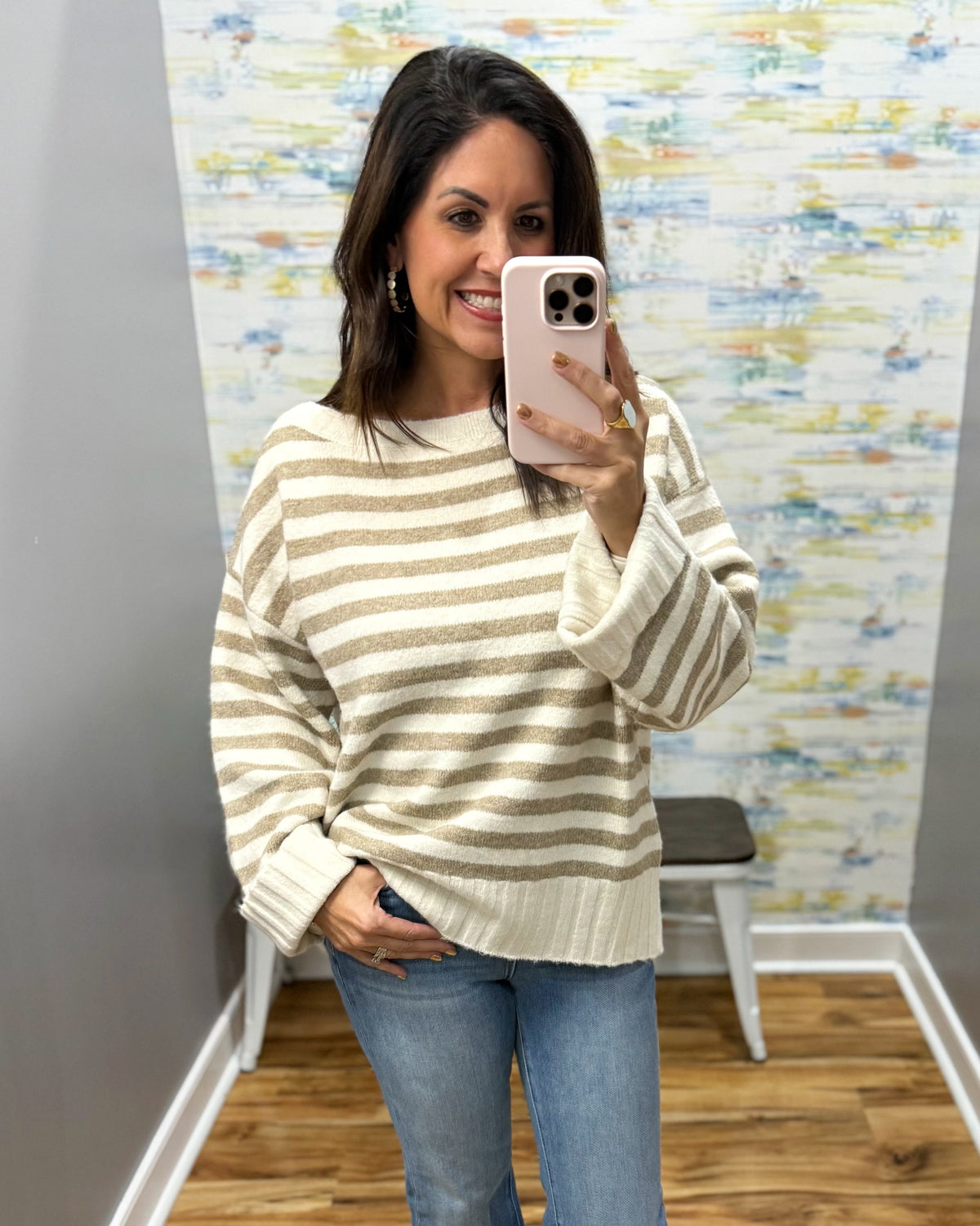 A woman takes a mirror selfie wearing a cream and tan striped knit sweater, blue jeans, and a light pink phone case in a fitting room with a patterned wallpaper background.