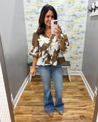 A woman takes a mirror selfie in a fitting room wearing a brown and white floral print blouse with ruffled sleeves, blue flare jeans, and tan pointed-toe flats.