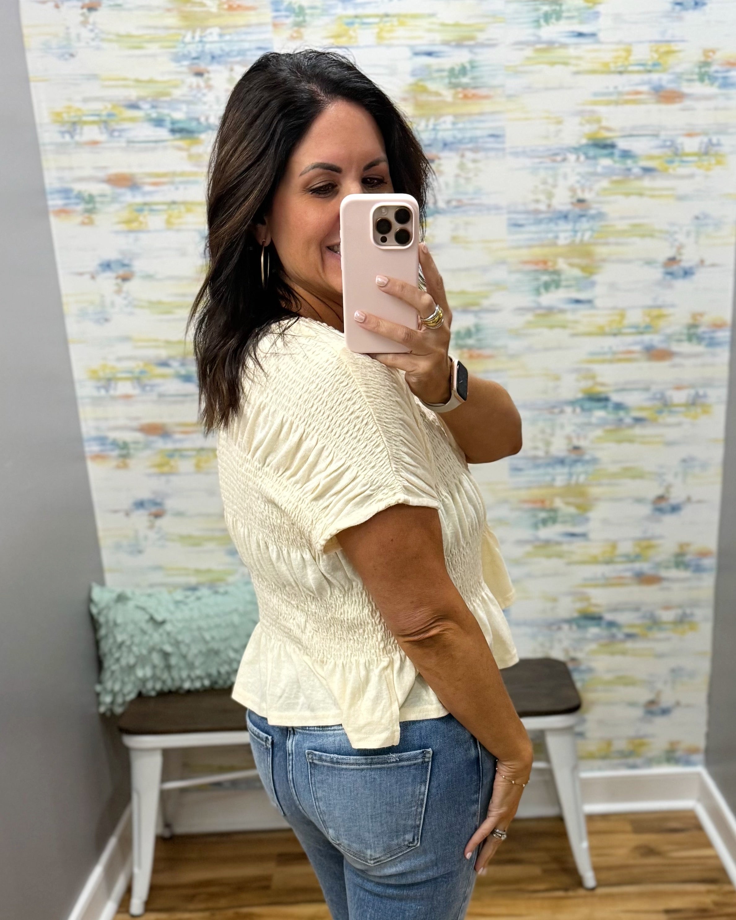 A woman takes a mirror selfie wearing a cream-colored, short-sleeved textured top and blue jeans in a fitting room with a patterned wall and a bench.