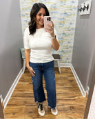 A woman takes a mirror selfie in a fitting room wearing a fitted white short-sleeve top, wide-leg blue jeans, and beige sneakers.