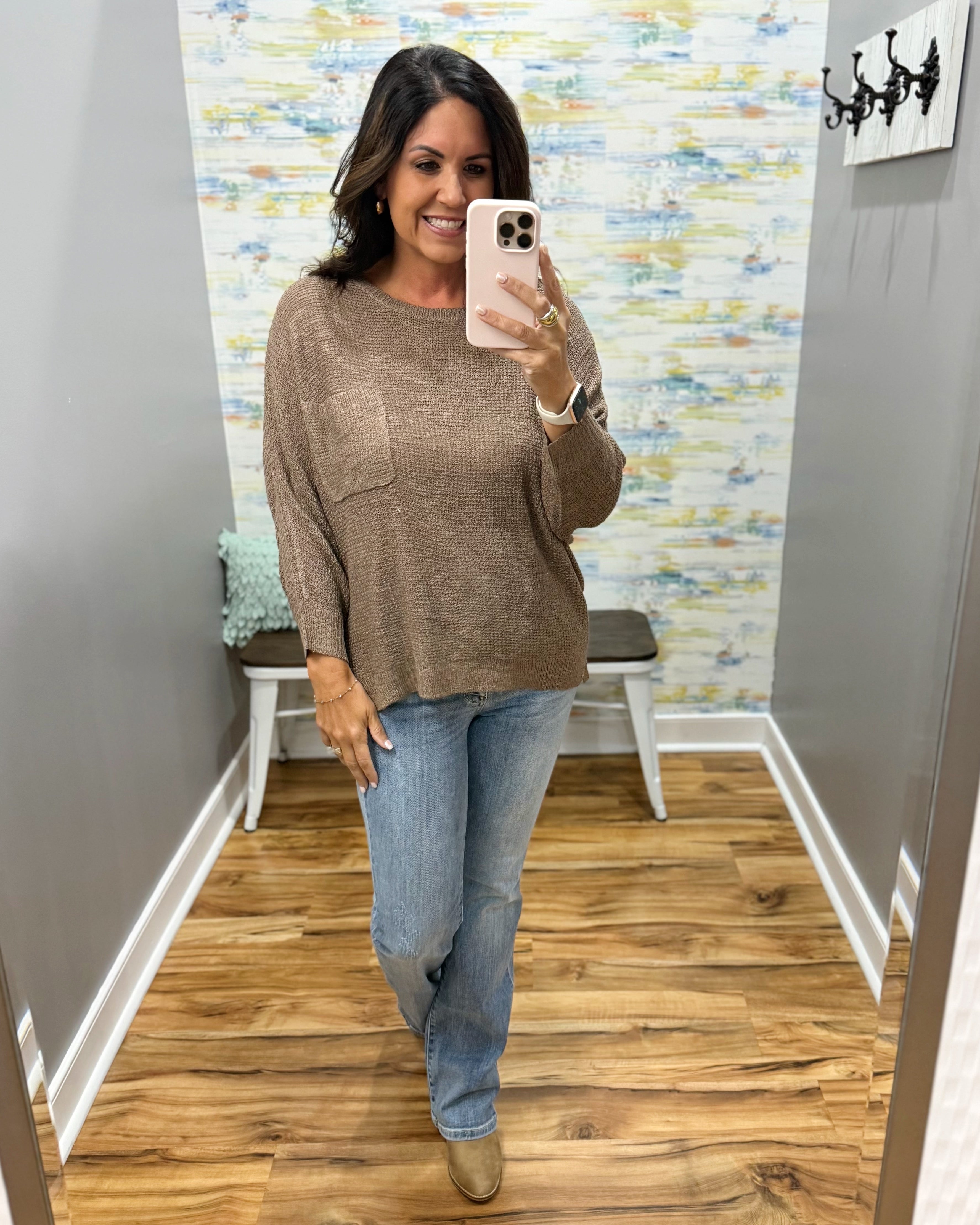 A woman takes a mirror selfie in a fitting room wearing a brown knit long-sleeve top with a chest pocket, blue jeans, and tan boots.