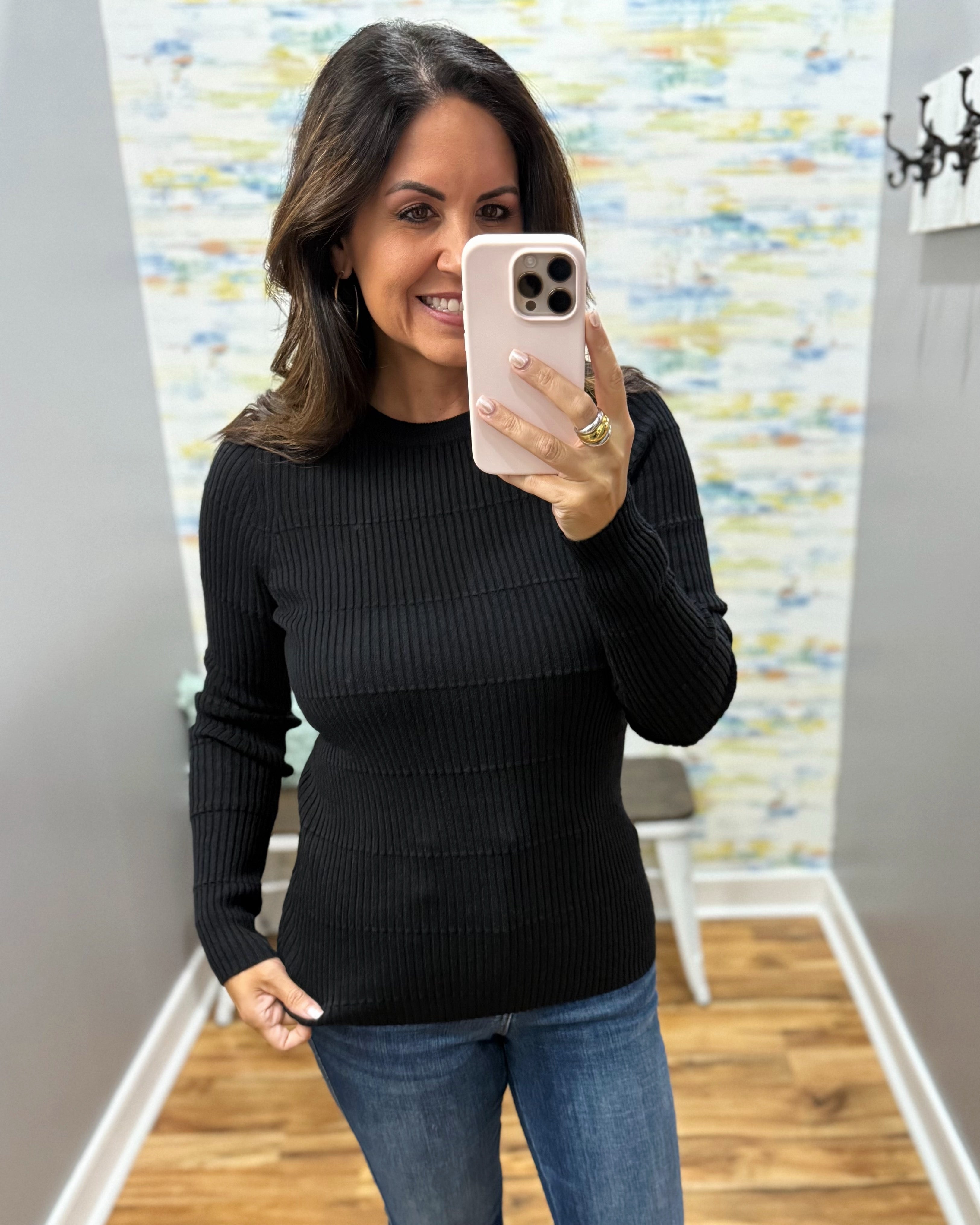 A woman taking a mirror selfie wearing a black ribbed long-sleeve top and blue jeans, holding a light pink smartphone.