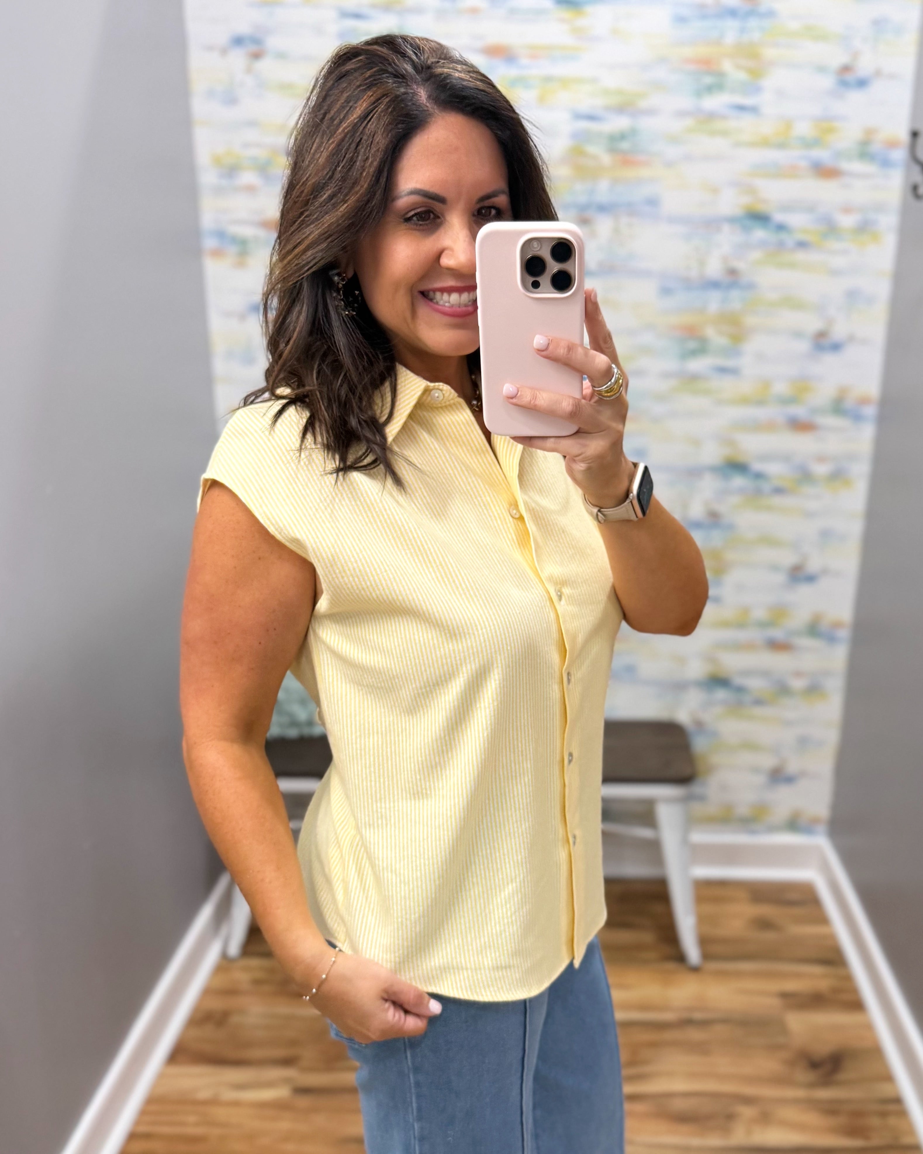 Woman taking a mirror selfie wearing a yellow shirt and blue jeans, holding a pink phone.