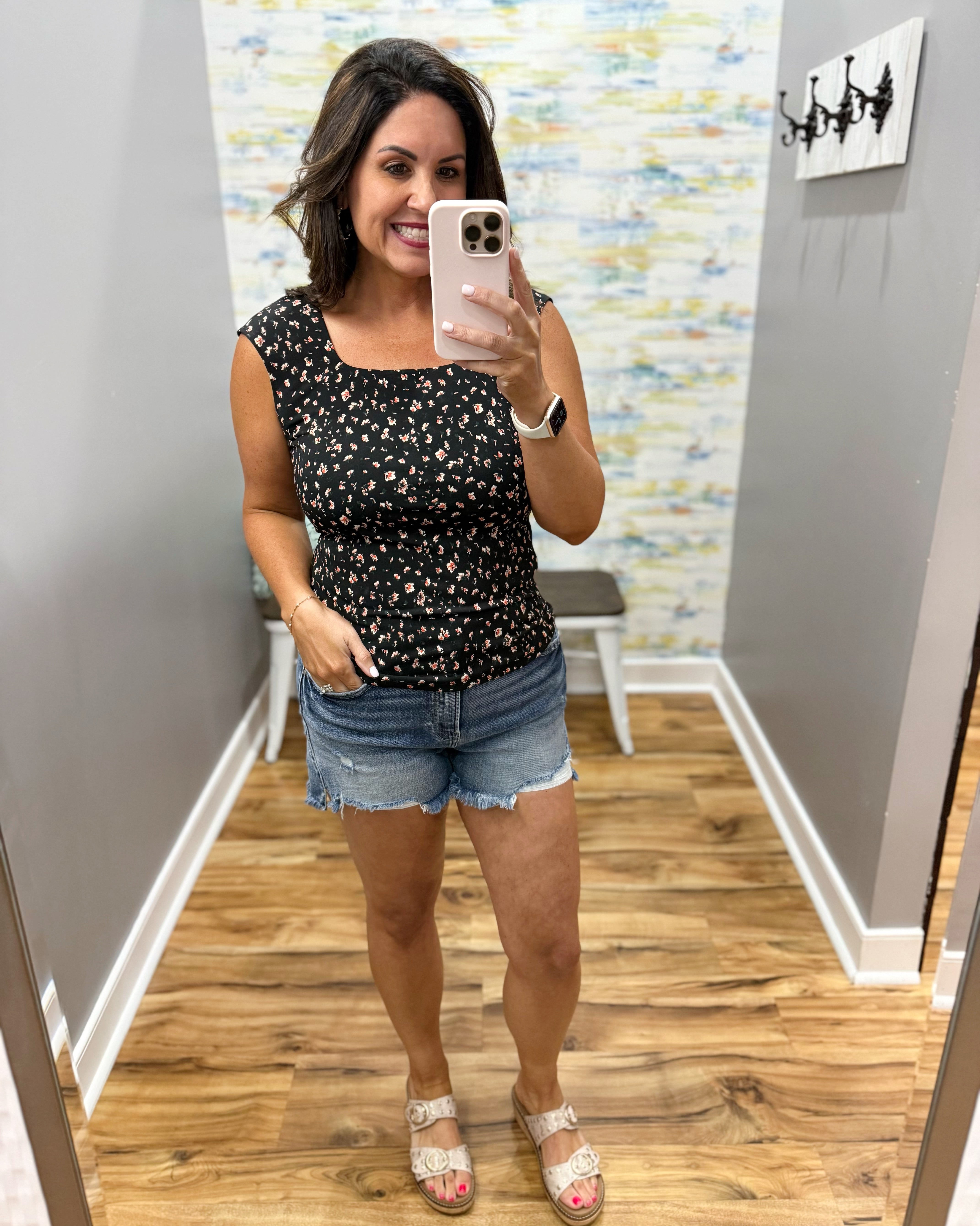 Woman taking a mirror selfie wearing a black floral top and denim shorts in a room with a patterned wall.