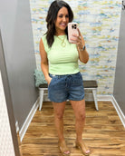 Woman wearing a light green sleeveless top and denim shorts in a room with a colorful wall.