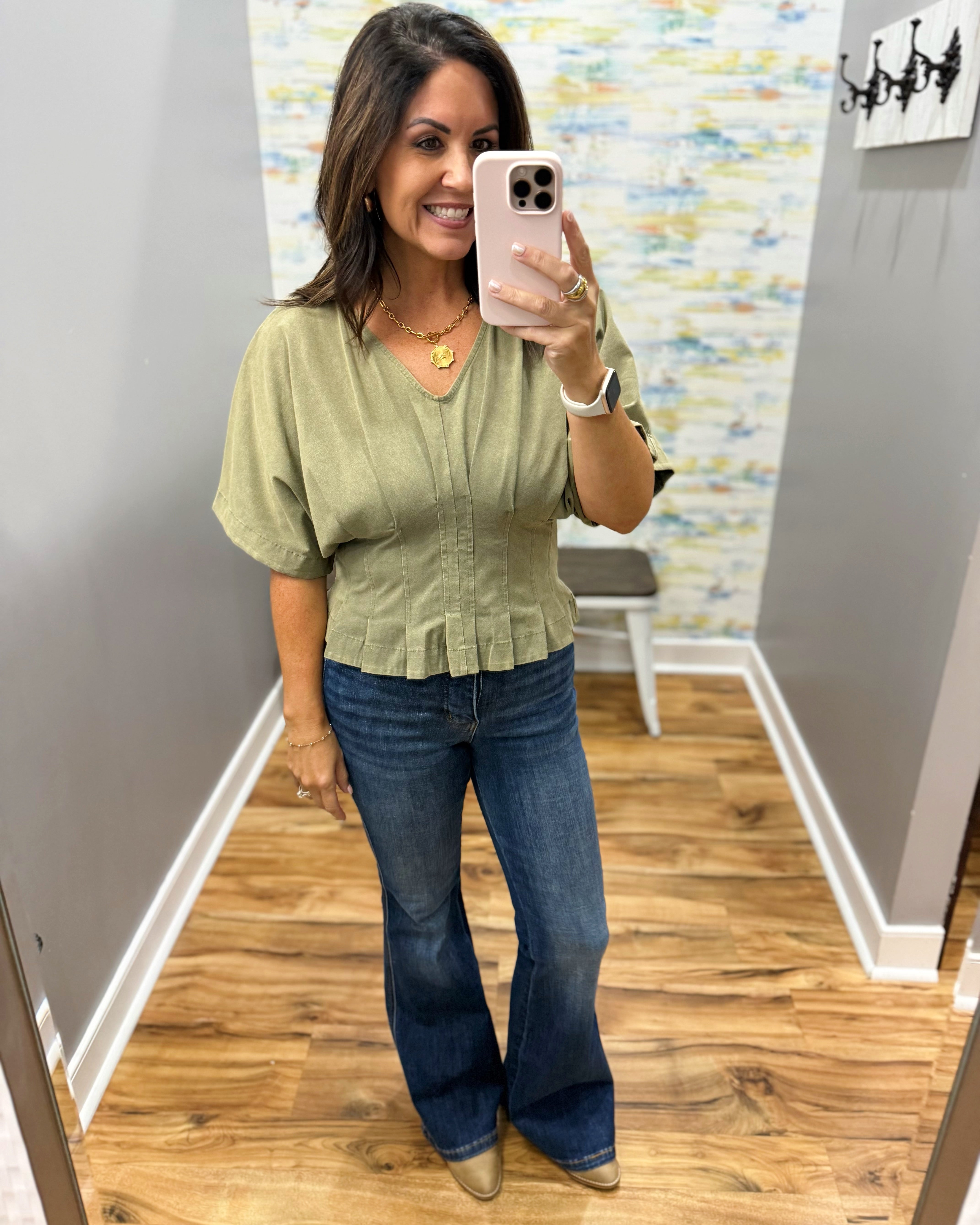 A woman takes a mirror selfie in a boutique showing an olive green short-sleeve blouse with a ruffled hem and blue flare jeans.