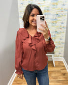 A woman takes a mirror selfie wearing a rust-colored long-sleeve blouse with ruffle details and blue jeans in a home setting.