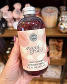 A hand holds a bottle of Sweet Grace Pink Flower Diffuser Refill in a shop setting with blurred candles in the background.