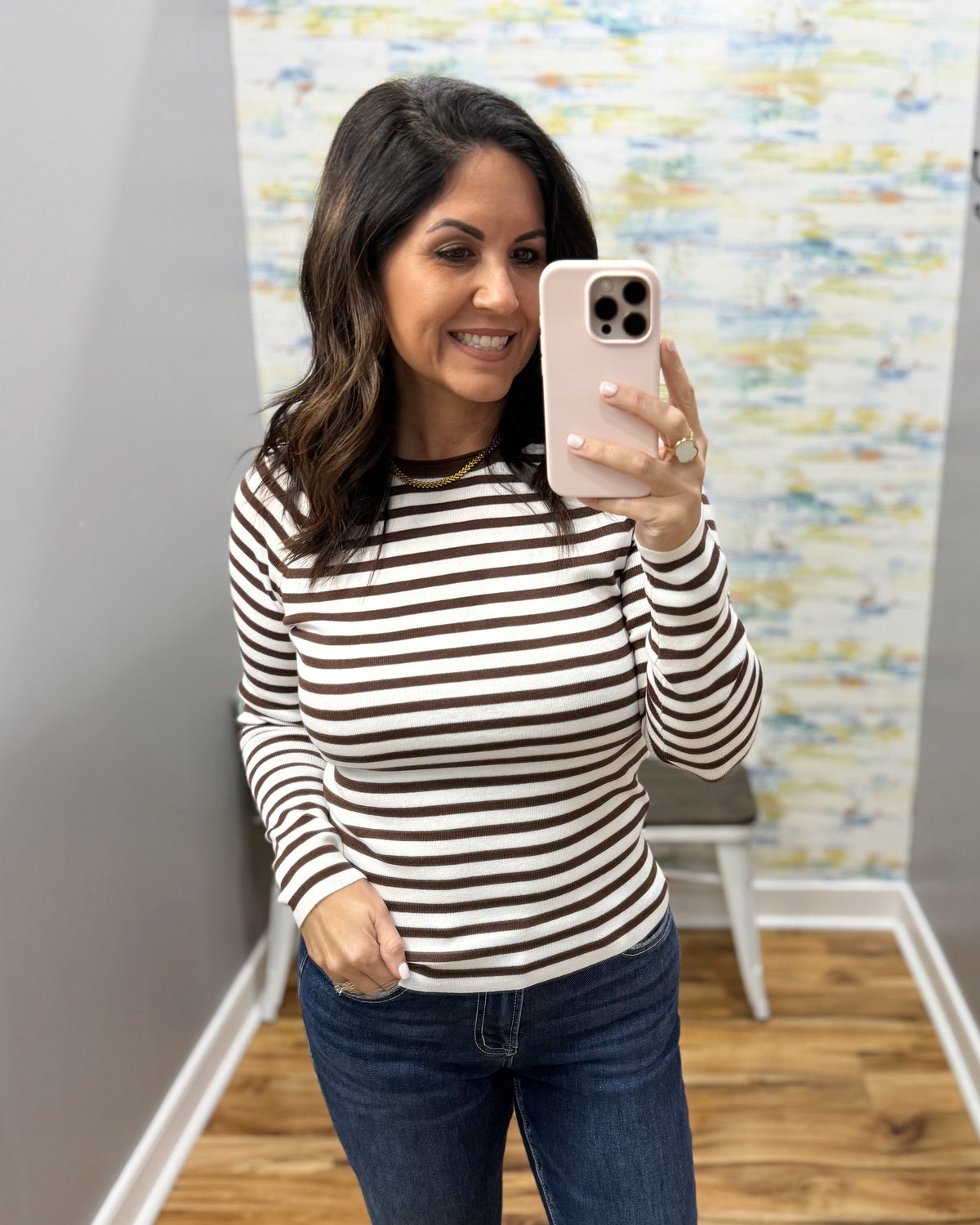 A woman taking a mirror selfie in a fitting room wearing a brown and white horizontally striped long-sleeve top and dark blue jeans.