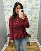 A woman taking a mirror selfie in a fitting room wearing a long-sleeve, smocked, and ruffled burgundy top with blue jeans.