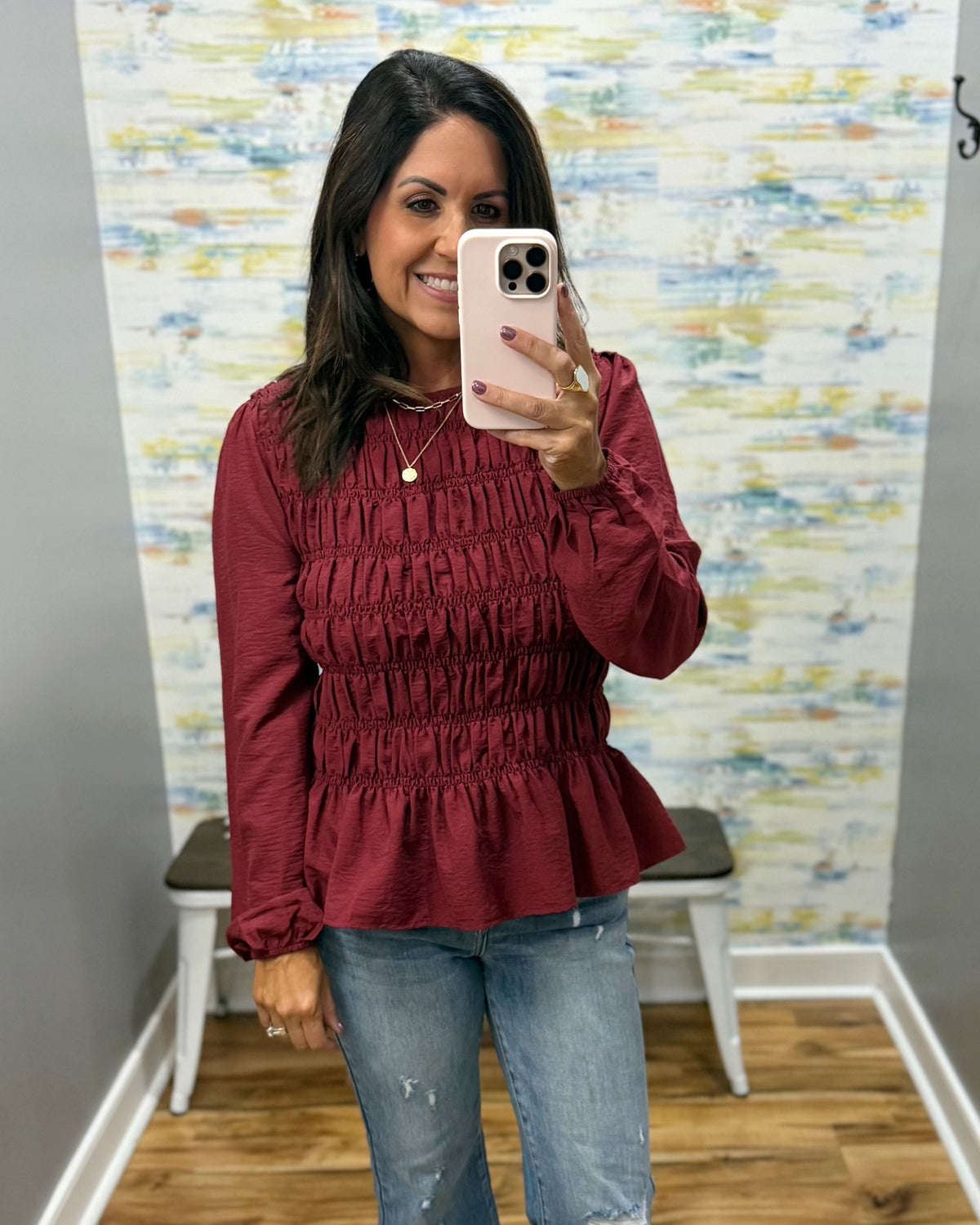 A woman taking a mirror selfie in a fitting room wearing a long-sleeve, smocked, and ruffled burgundy top with blue jeans.