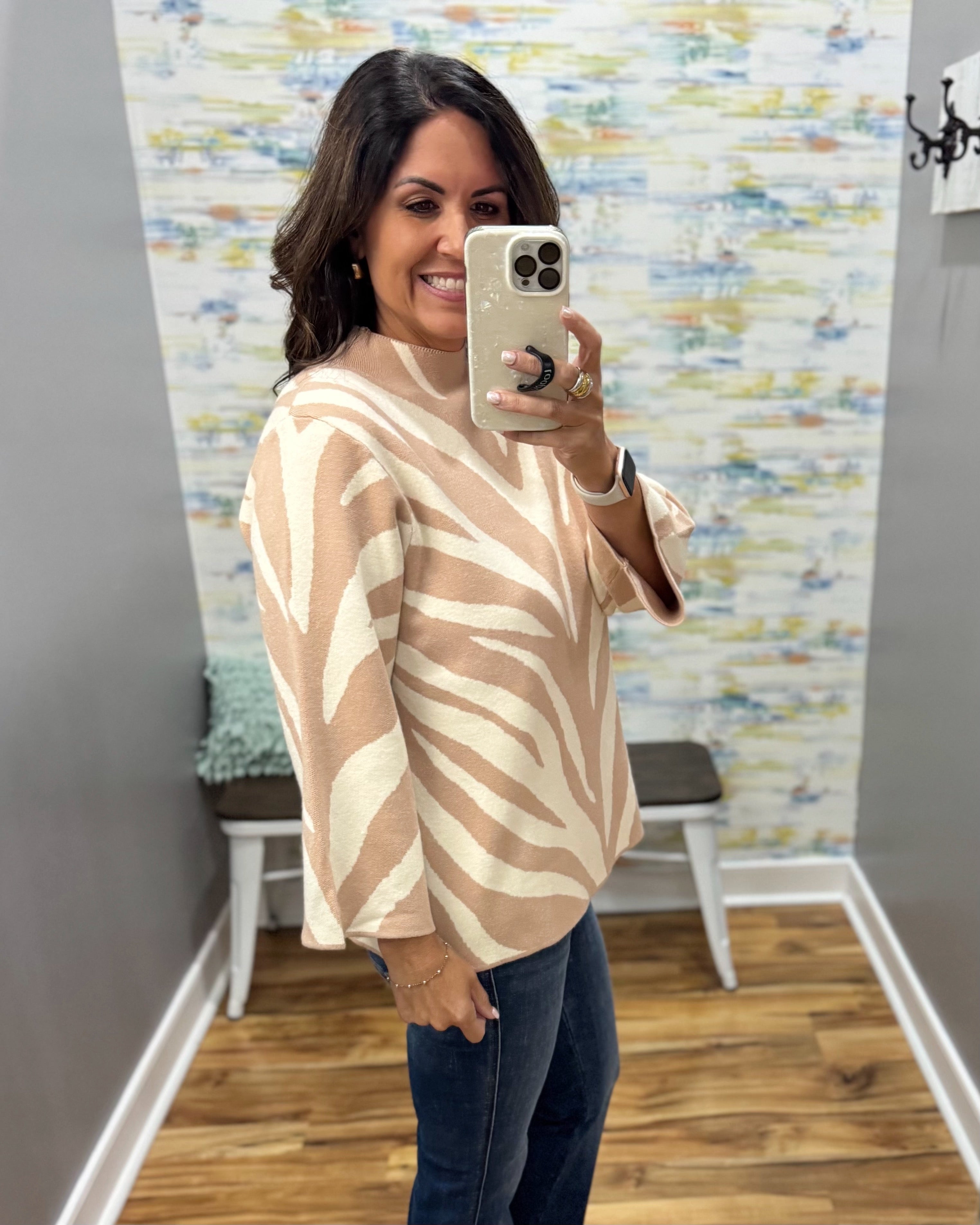 A woman takes a mirror selfie in a fitting room wearing a beige and cream zebra-print knit sweater and dark blue jeans.