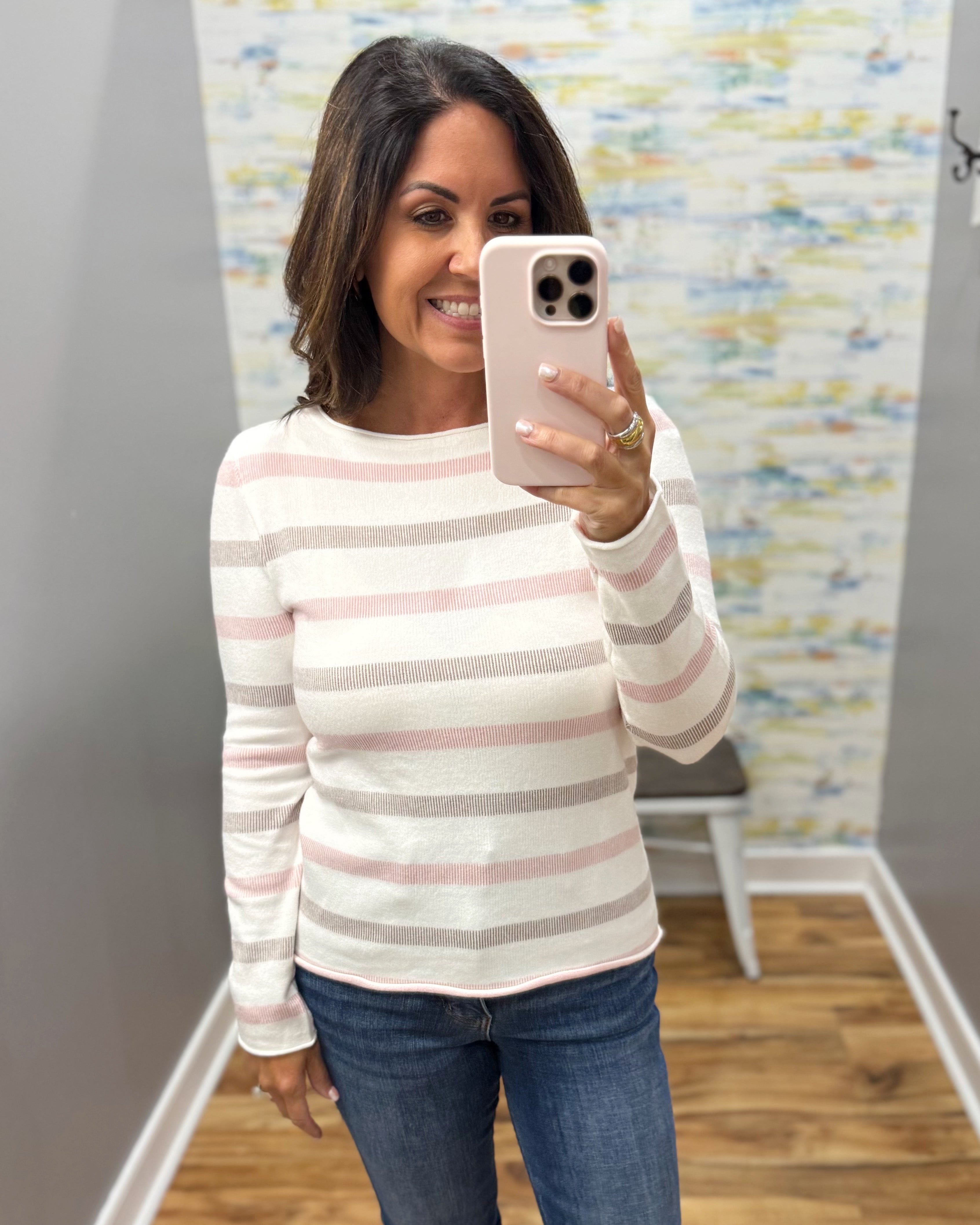 A woman takes a mirror selfie wearing a long-sleeve knit sweater with horizontal stripes in cream, light pink, and taupe, paired with blue jeans.