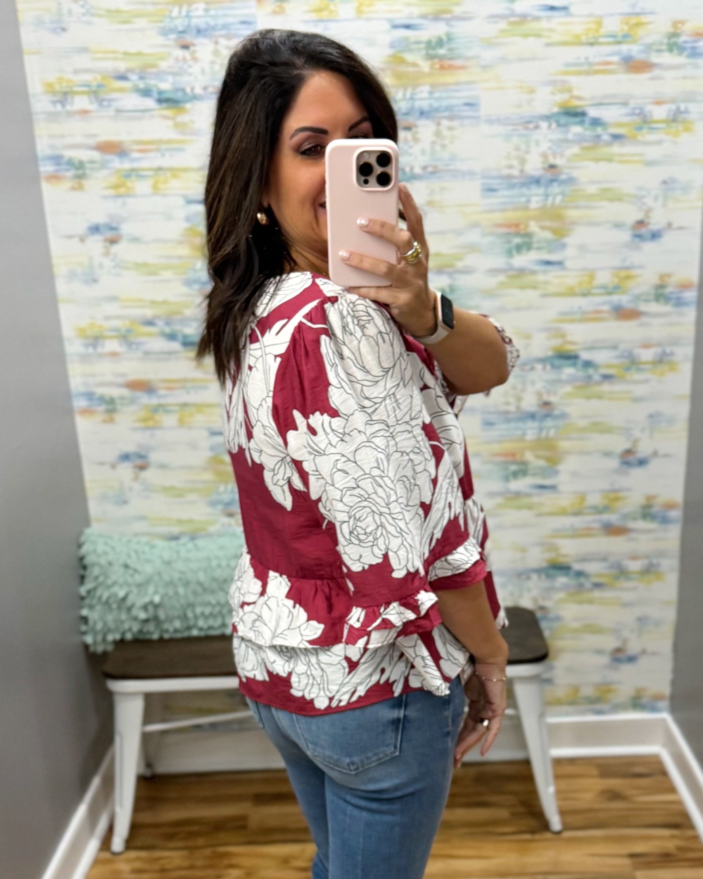 A woman takes a mirror selfie wearing a red and white floral print blouse with blue jeans, standing in front of a patterned wallpaper background.