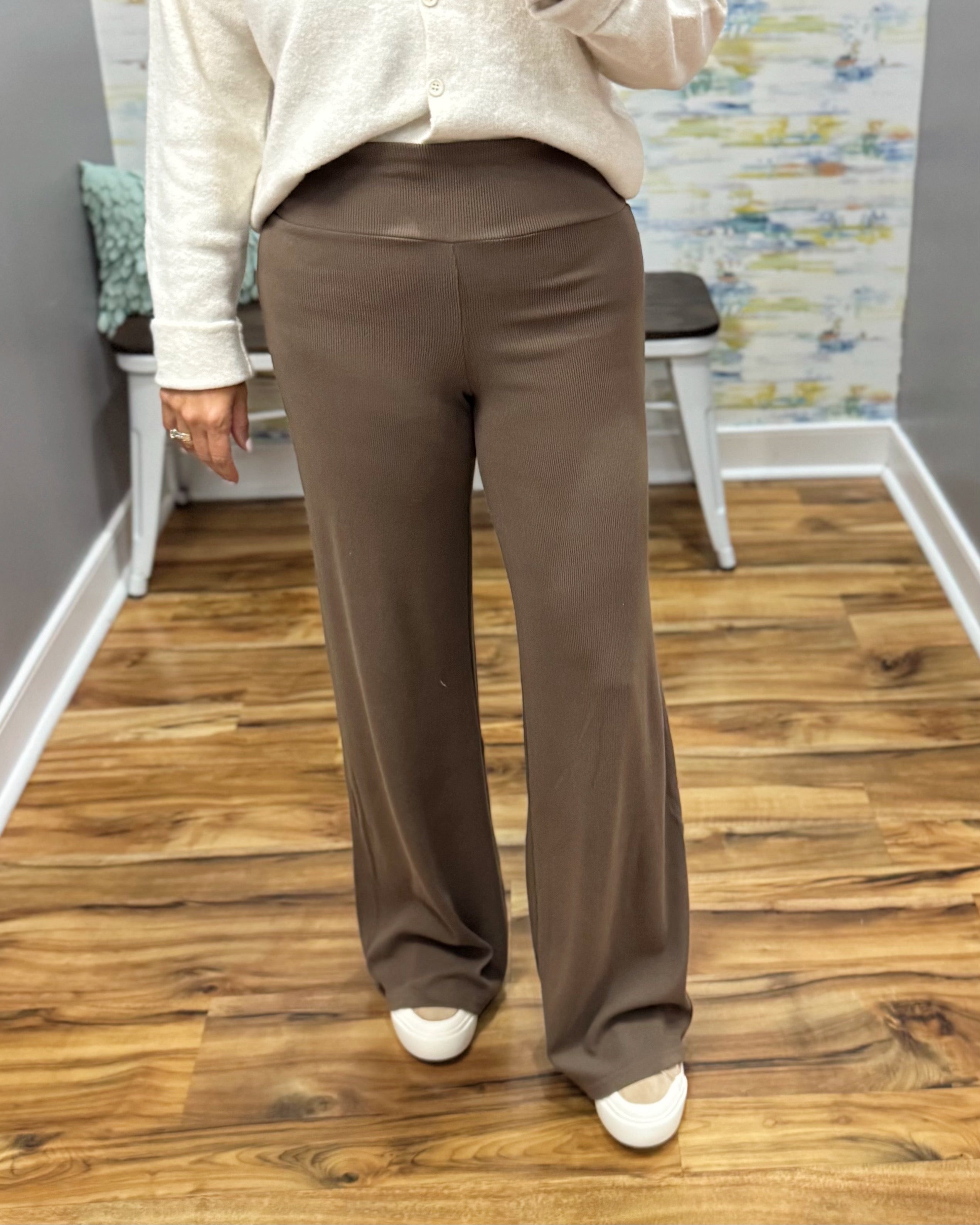 A person wearing brown wide-leg pants, a cream knit cardigan, and white shoes, standing on a wood floor in an indoor room with a bench and abstract wall art in the background.