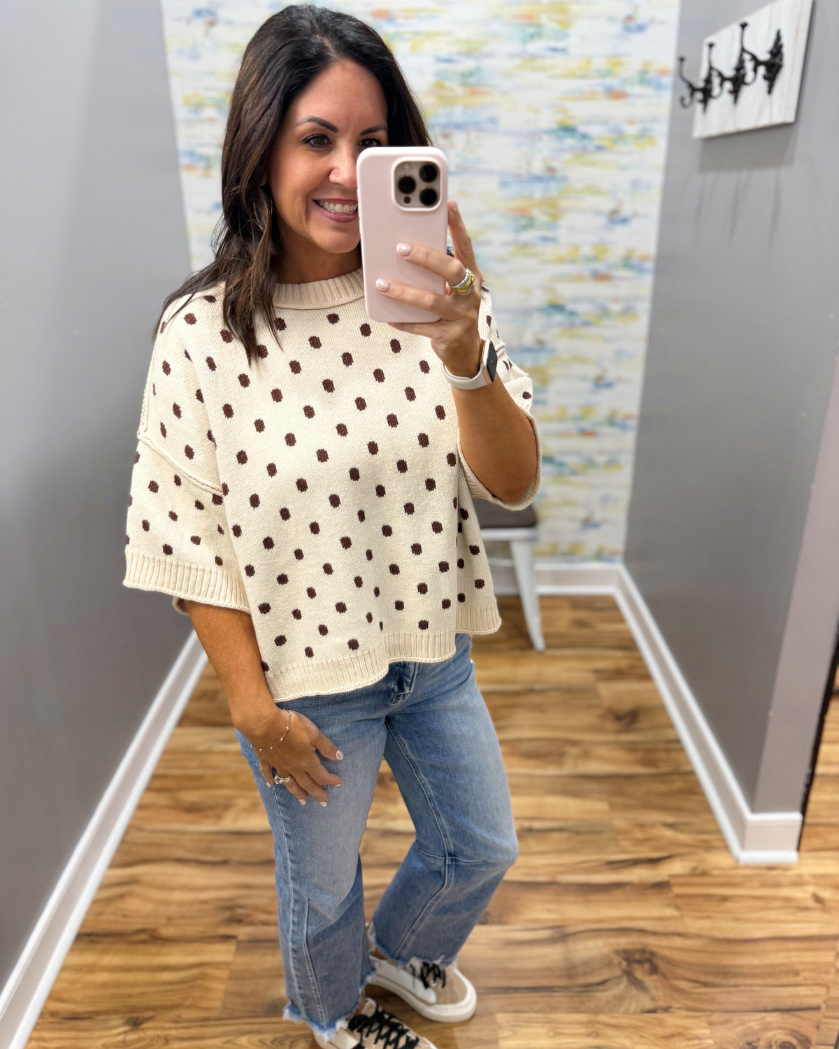 A woman takes a mirror selfie wearing a cream knit sweater with brown polka dots, paired with light blue jeans and white sneakers, in an indoor hallway.