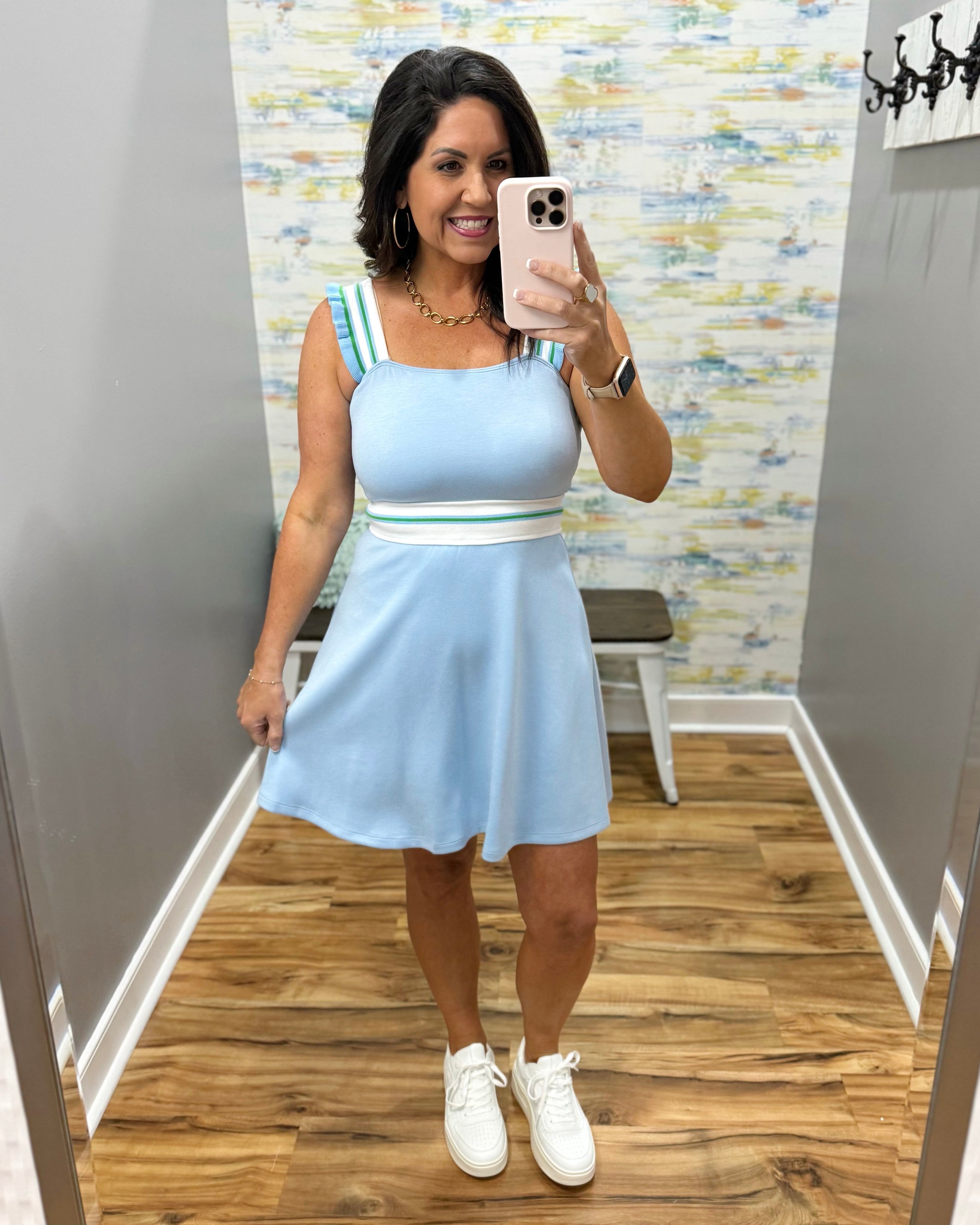 Woman taking a mirror selfie wearing a light blue dress with white sneakers in a room with a patterned wall.