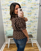 A woman takes a mirror selfie wearing a brown long-sleeve blouse with white polka dots and blue jeans in a fitting room with a patterned wallpaper background.