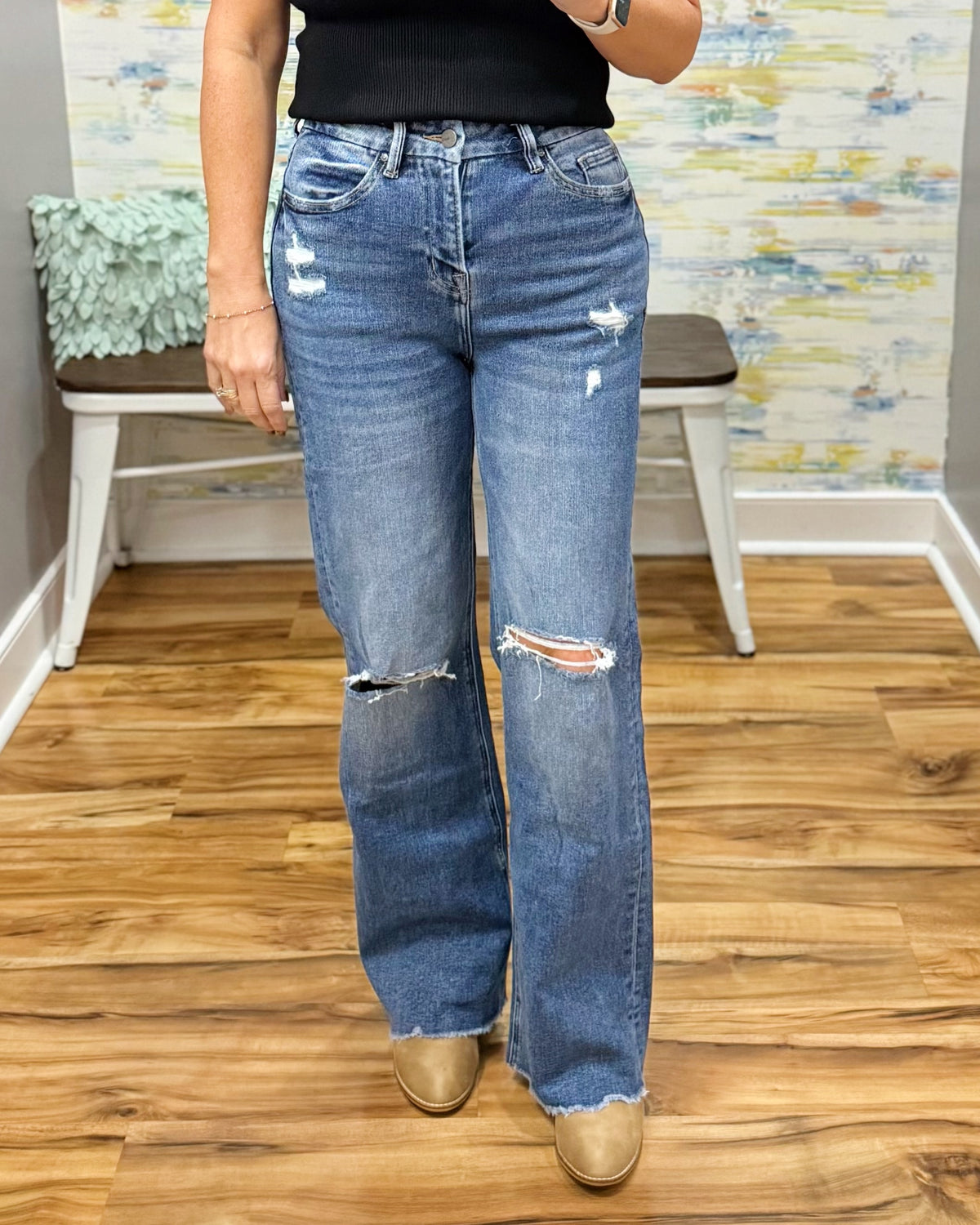 A person wearing mica wide leg distressed jeans with ripped details on the thighs and knees, paired with a black top and tan boots, standing in an indoor room with wood floors and a bench.