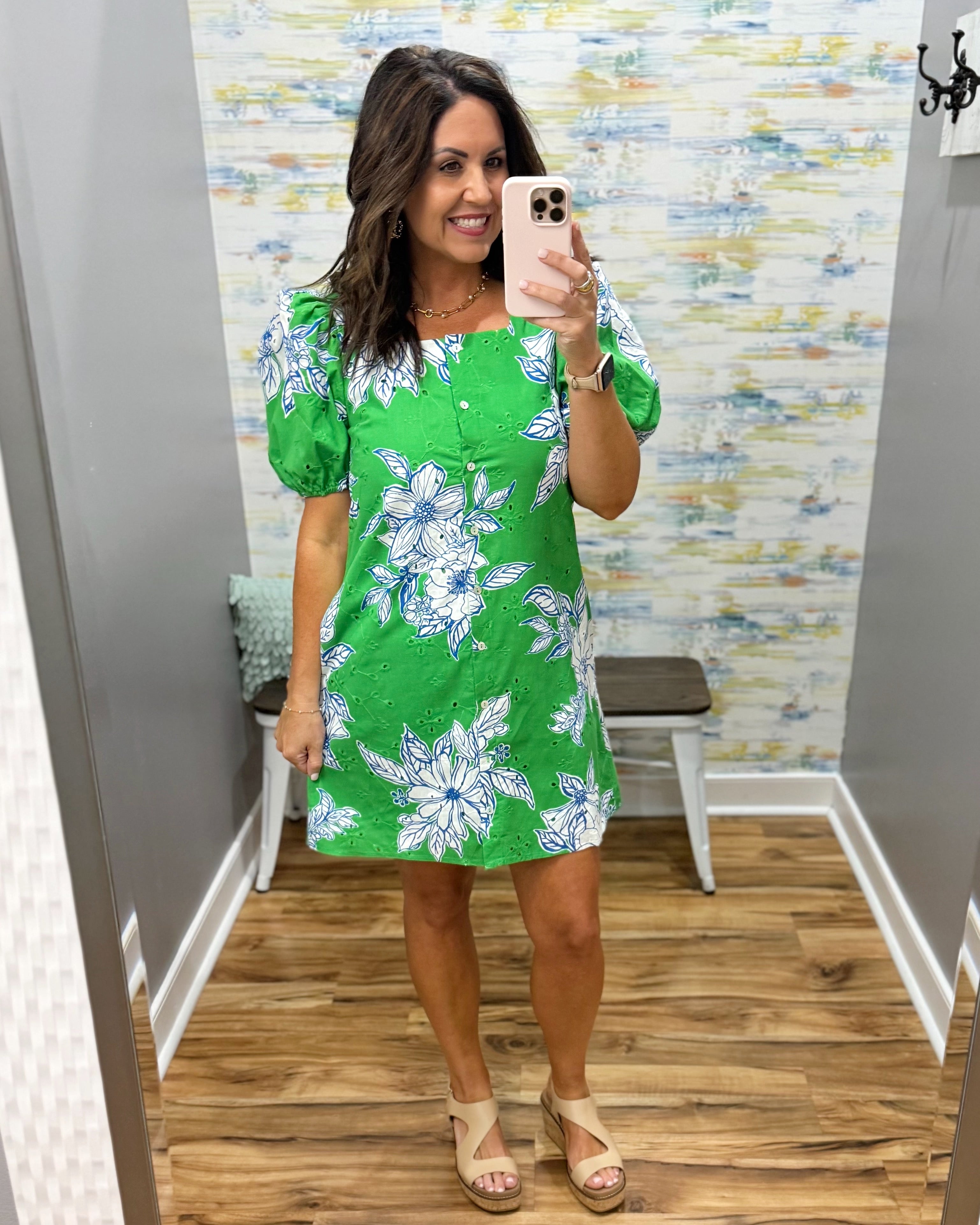Woman taking a mirror selfie wearing a green floral dress in a room with patterned wallpaper.