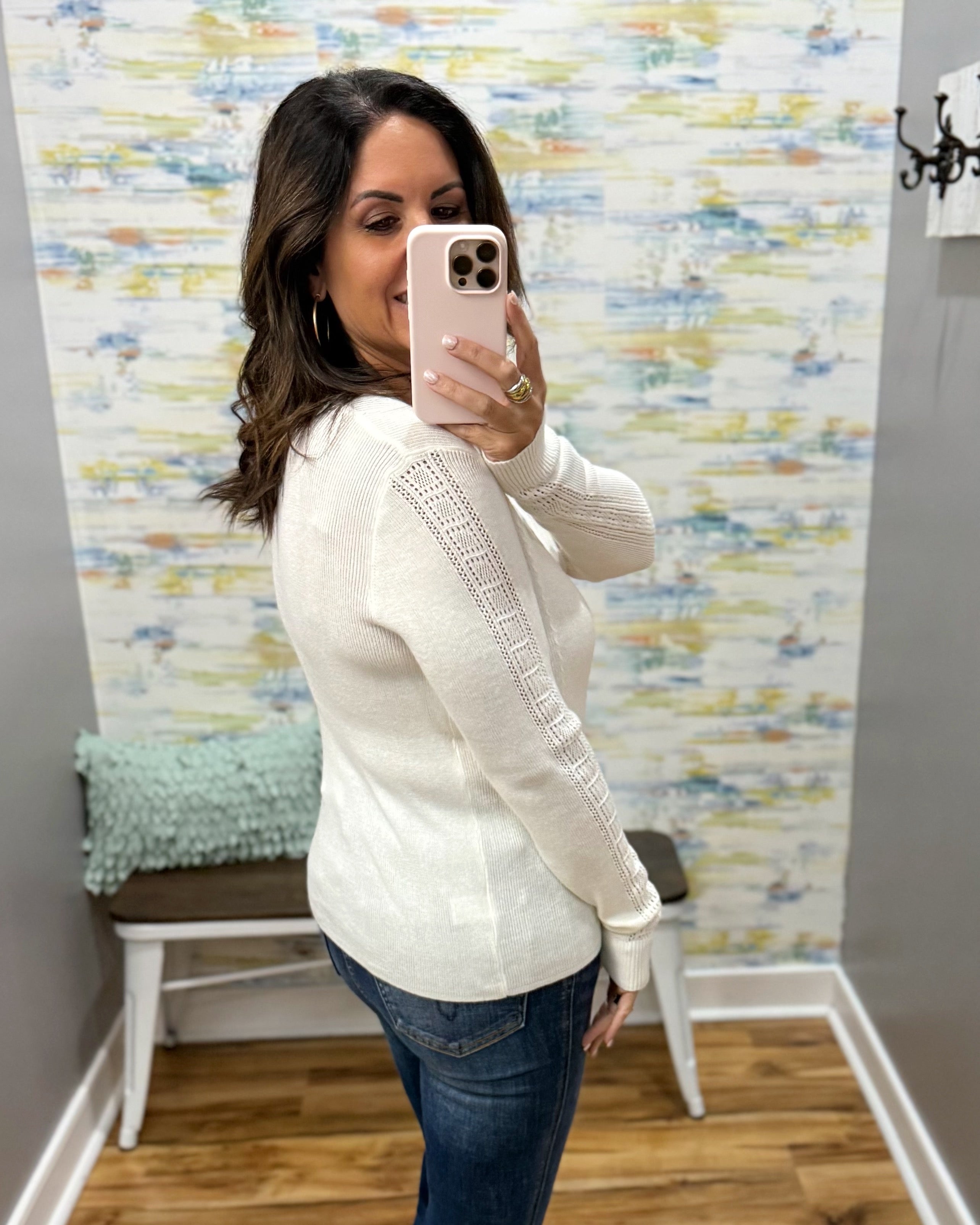 A woman takes a mirror selfie in a fitting room wearing a cream-colored knit sweater with textured lace-like sleeve panels and blue jeans.