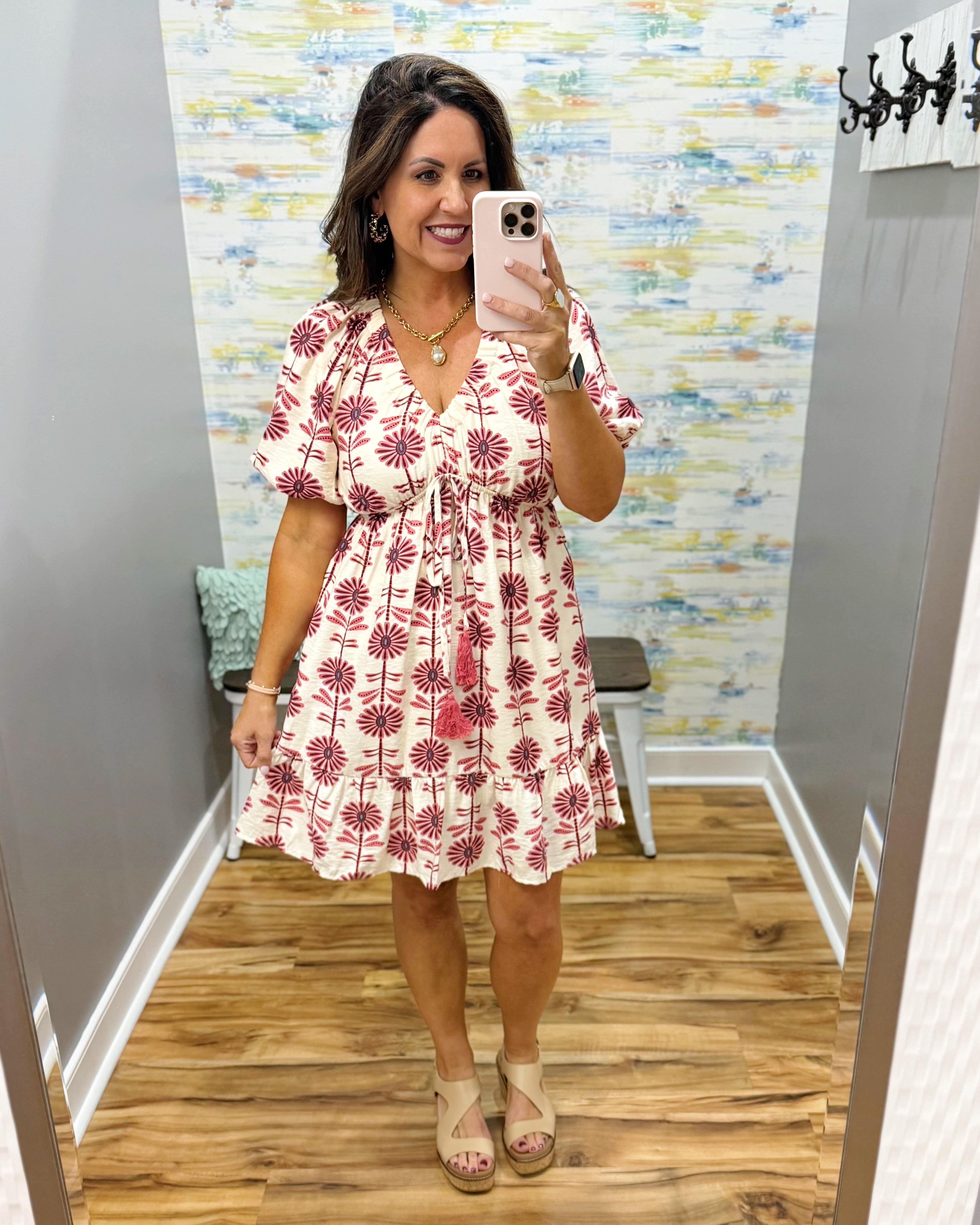 Woman taking a mirror selfie wearing a white dress with red floral pattern in a room with wooden flooring and colorful wall.