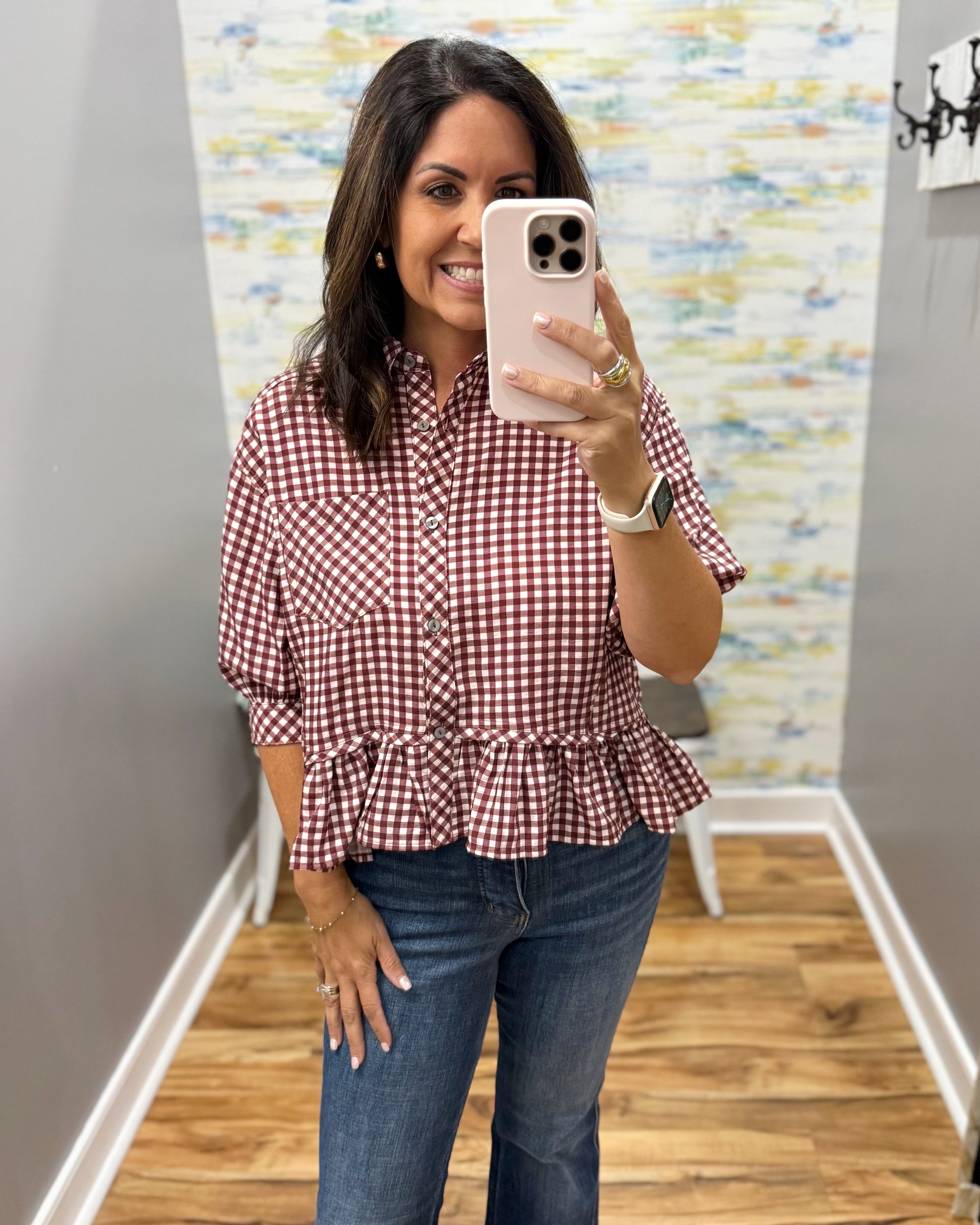 A woman takes a mirror selfie wearing a red and white gingham button-up blouse with a ruffled hem and blue jeans in a home setting with wood floors and a patterned wallpaper background.
