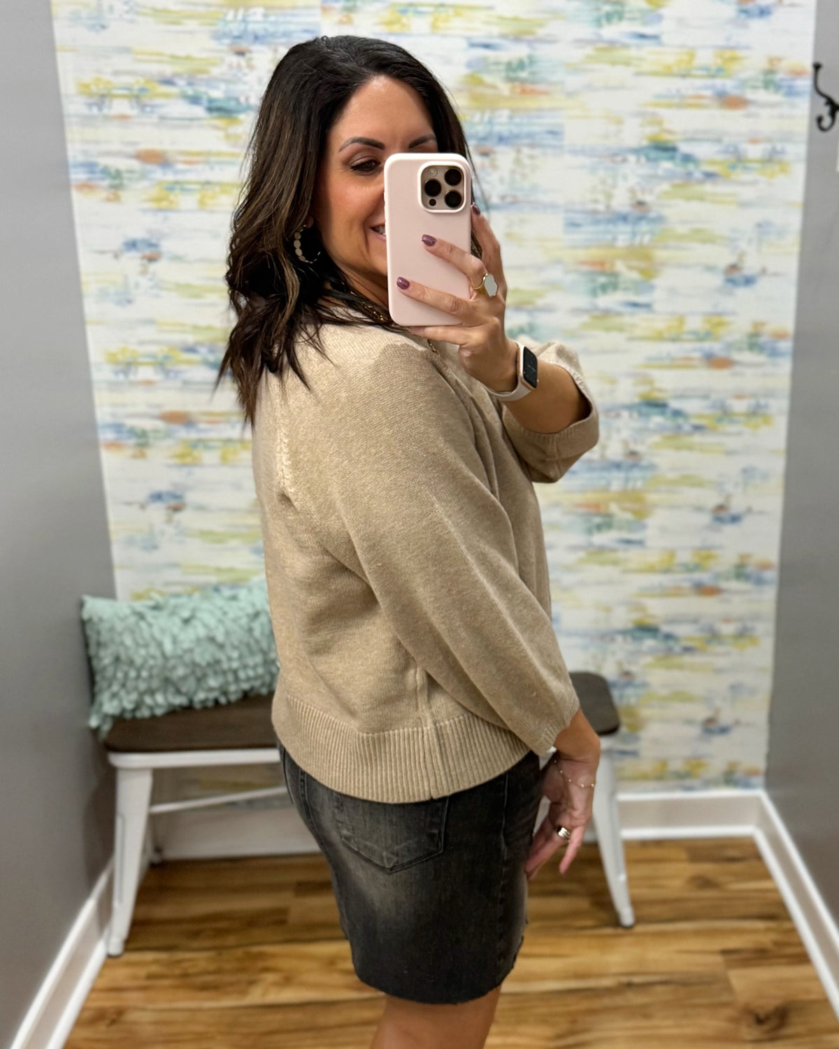 A woman takes a mirror selfie in a fitting room wearing a beige knit sweater and a dark gray denim skirt. The background features patterned wallpaper, a wooden floor, and a small bench with a mint green pillow.