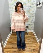 A woman takes a mirror selfie in a fitting room wearing a pink and white striped knit sweater, blue flare jeans, and brown boots.
