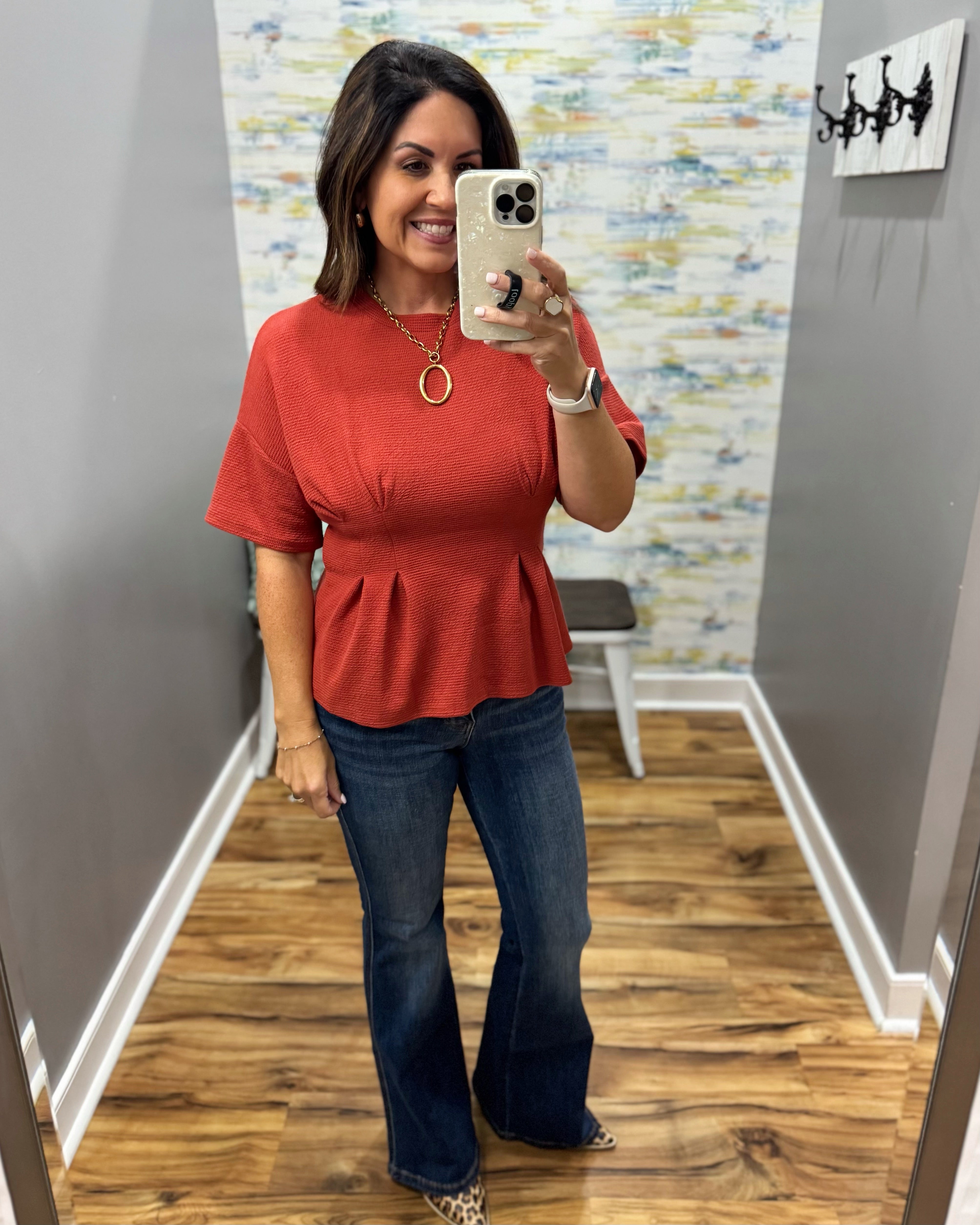 A woman takes a mirror selfie wearing a terracotta orange short-sleeve peplum top and dark blue flare jeans in a fitting room with wood floors and gray walls.