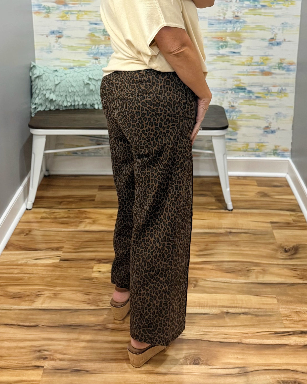 A person wearing brown leopard-print wide-leg pants, a cream short-sleeve top, and wedge sandals, standing in a room with wood floors and a bench in the background.