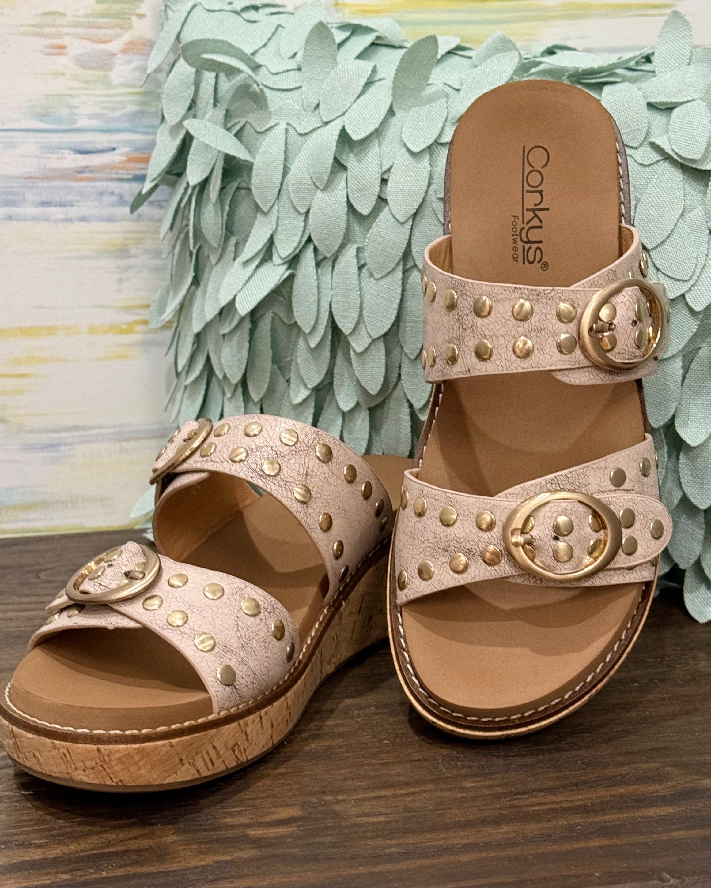 Pair of beige sandals with gold embellishments on a wooden surface with decorative green leaves in the background.