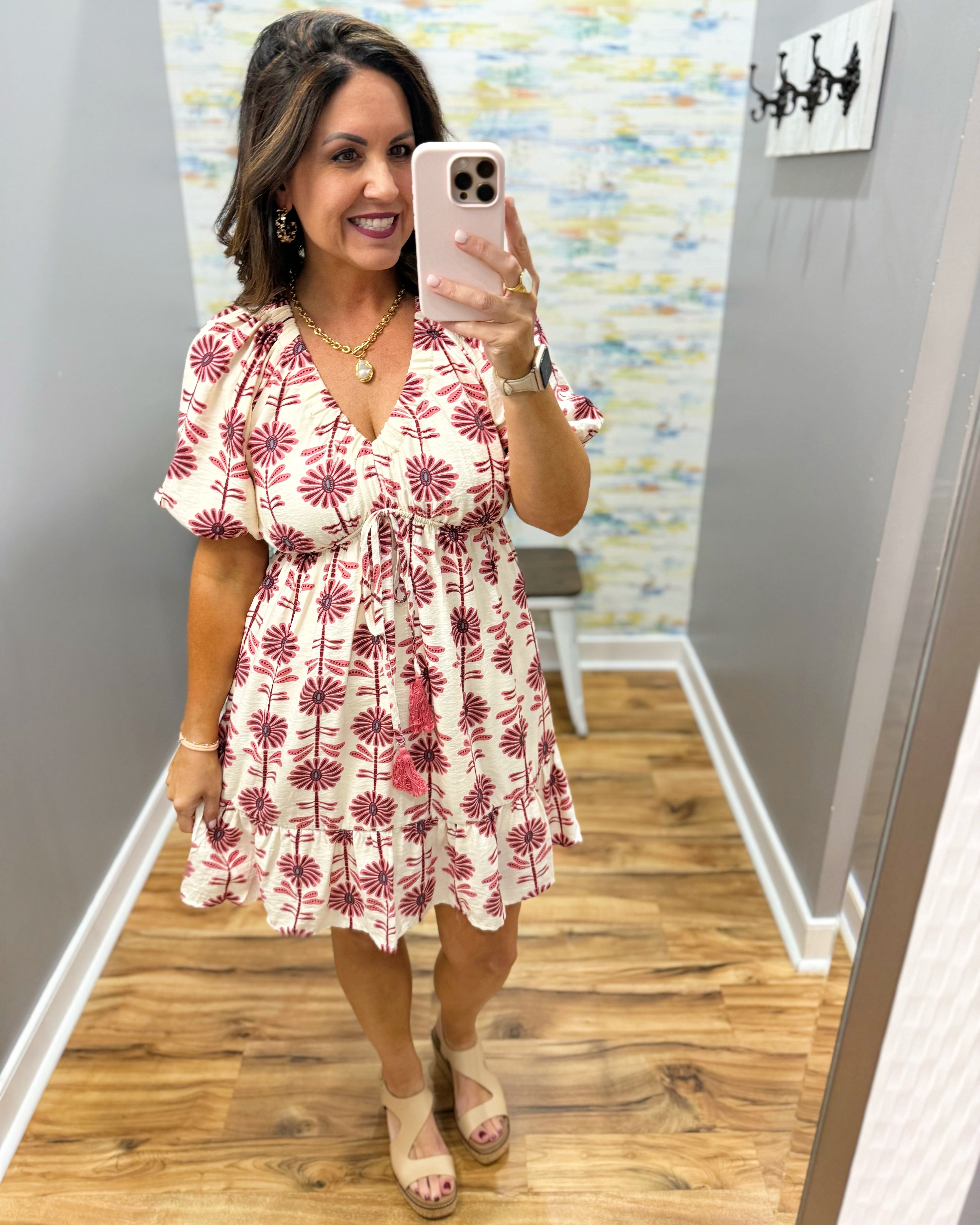 Woman taking a mirror selfie wearing a white dress with red floral pattern in a room with wooden flooring and colorful wall.