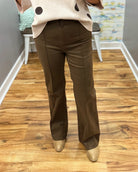 A person shown from the waist down wearing dark brown straight-leg trousers, a beige sweater with dark polka dots, and metallic gold shoes, standing on a wooden floor in an indoor room.
