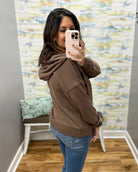 A woman takes a mirror selfie wearing a brown pullover hoodie and blue jeans in a room with a patterned wallpaper background.