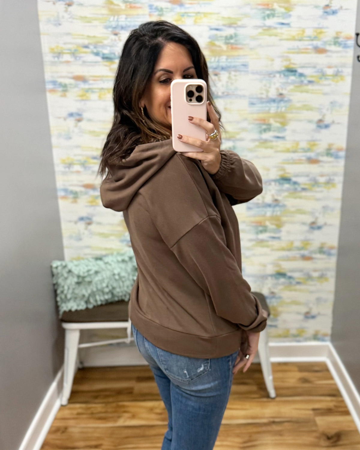 A woman takes a mirror selfie wearing a brown pullover hoodie and blue jeans in a room with a patterned wallpaper background.