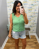 Woman taking a mirror selfie wearing a green sleeveless top and denim shorts in a room with a patterned wall.