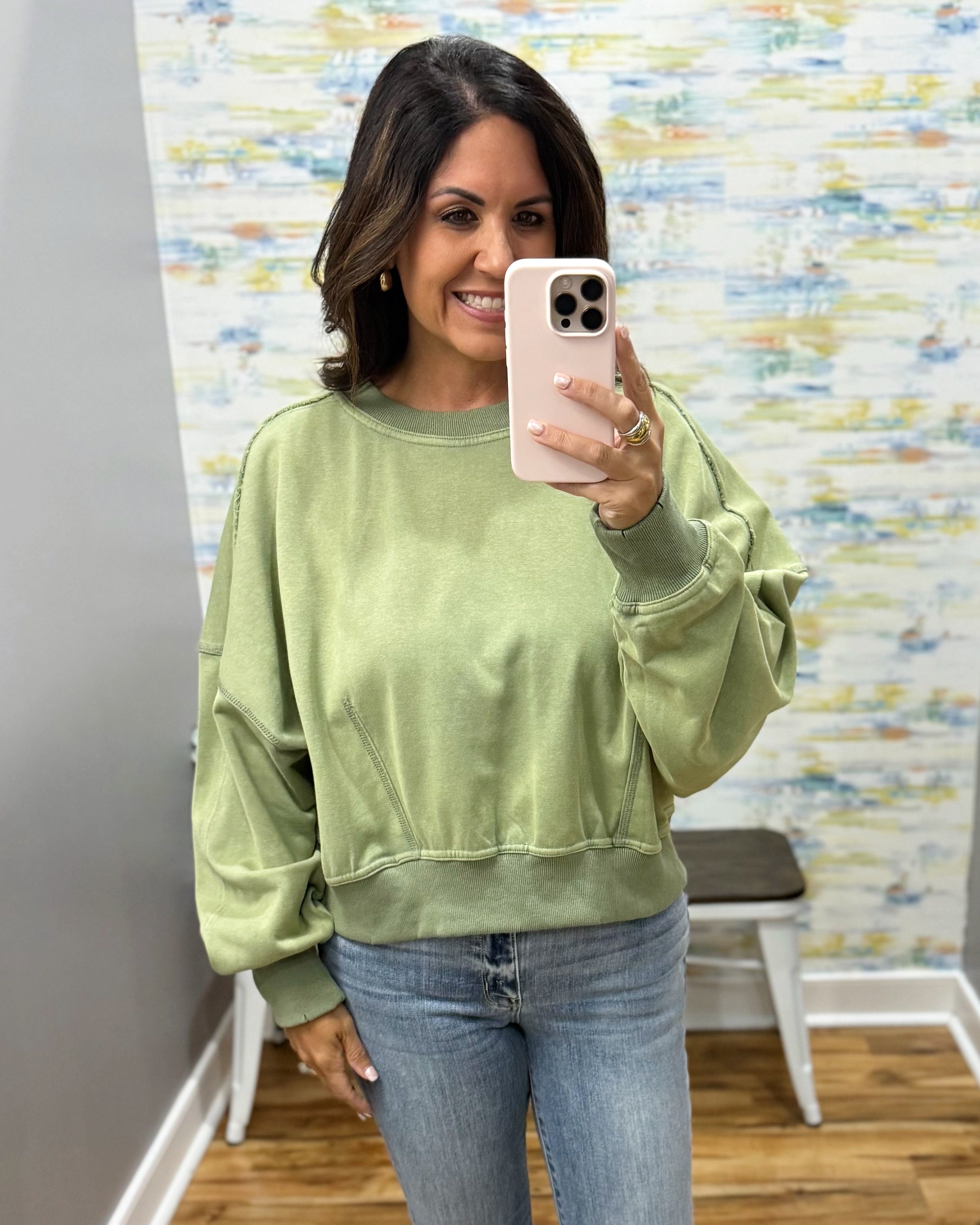 A woman takes a mirror selfie wearing a sage green crewneck sweatshirt and blue jeans in an indoor setting with a patterned wall and wooden floor.