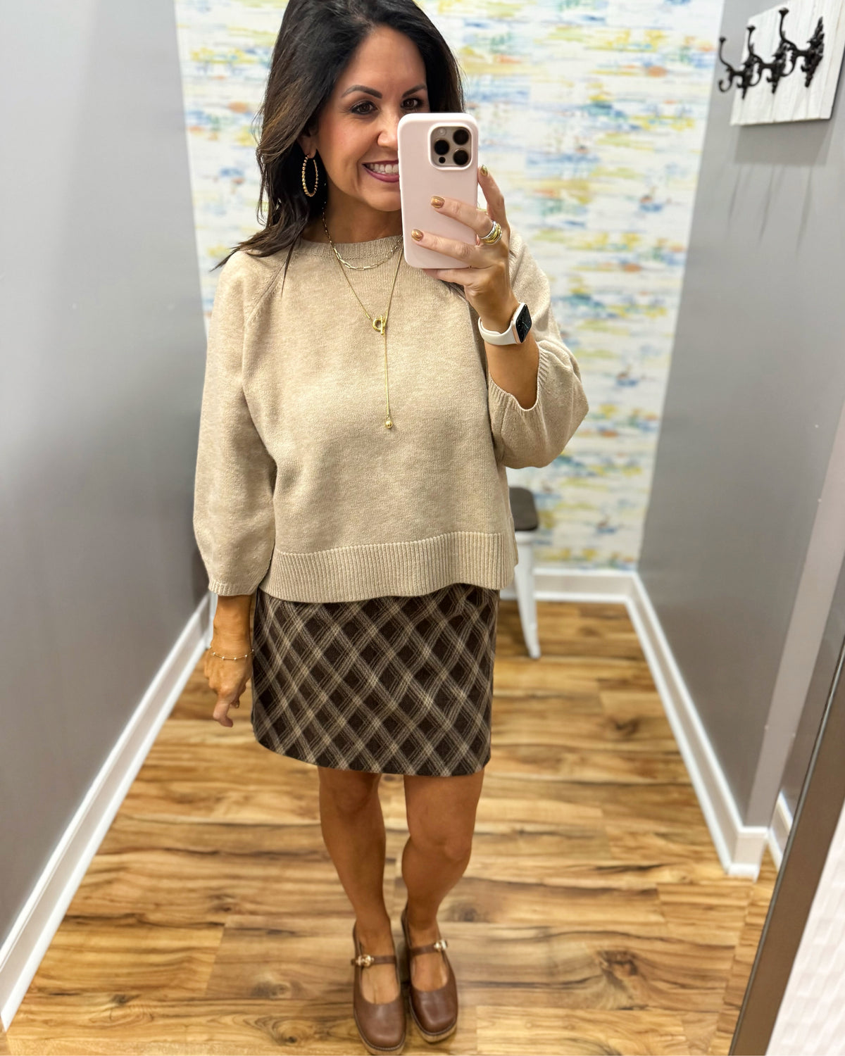 A woman takes a mirror selfie in a fitting room wearing a beige knit crewneck sweater, a brown and black plaid mini skirt, and Corkys Pity Party shoes in Cognac.