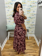 A woman takes a mirror selfie in a fitting room wearing a dark burgundy maxi dress with a pink floral pattern. The fitting room has a wood floor, a bench with a mint green pillow, and a colorful abstract wallpaper.