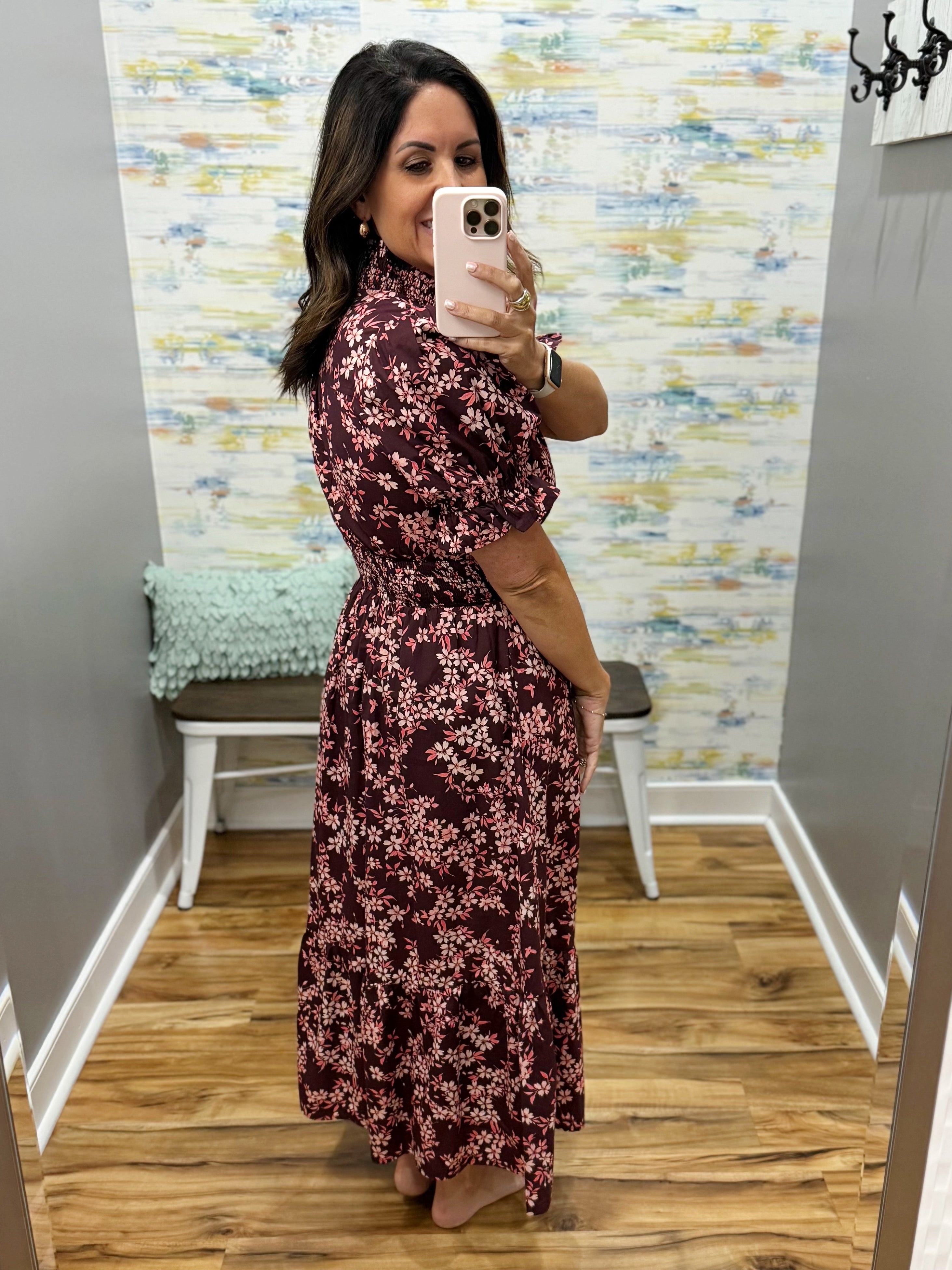A woman takes a mirror selfie in a fitting room wearing a dark burgundy maxi dress with a pink floral pattern. The fitting room has a wood floor, a bench with a mint green pillow, and a colorful abstract wallpaper.