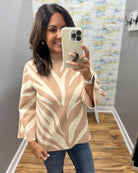 A woman takes a mirror selfie wearing a beige and cream abstract patterned knit top with flared bell sleeves, paired with blue jeans.