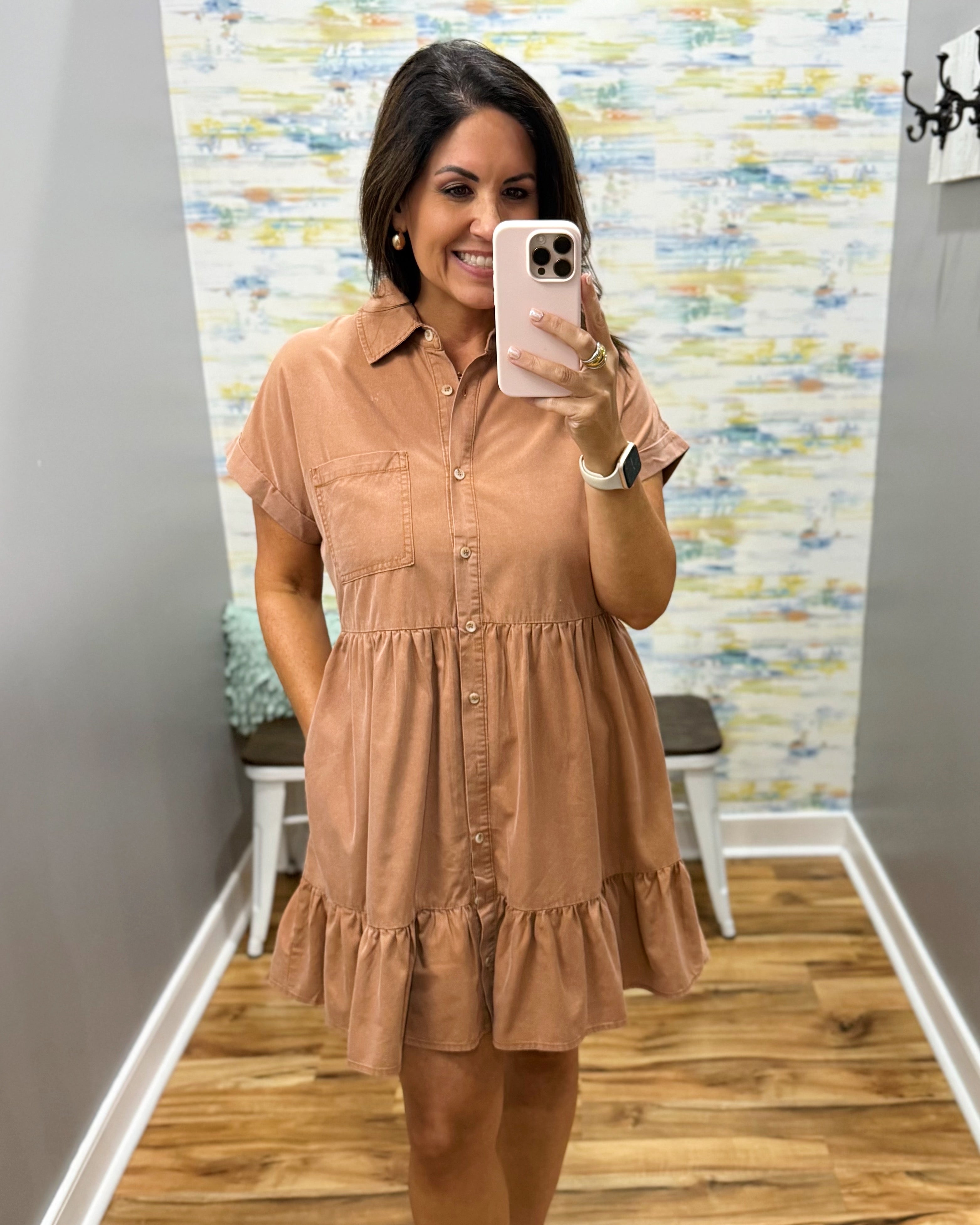 A woman takes a mirror selfie in a boutique wearing a short-sleeve, tiered button-up dress in a warm tan or camel color. The background features gray walls, a patterned wallpaper, and a wooden floor.