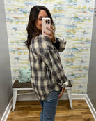 A woman takes a mirror selfie wearing an oversized plaid flannel shirt in beige, brown, and black, paired with blue jeans, in front of a decorative wall with a bench.