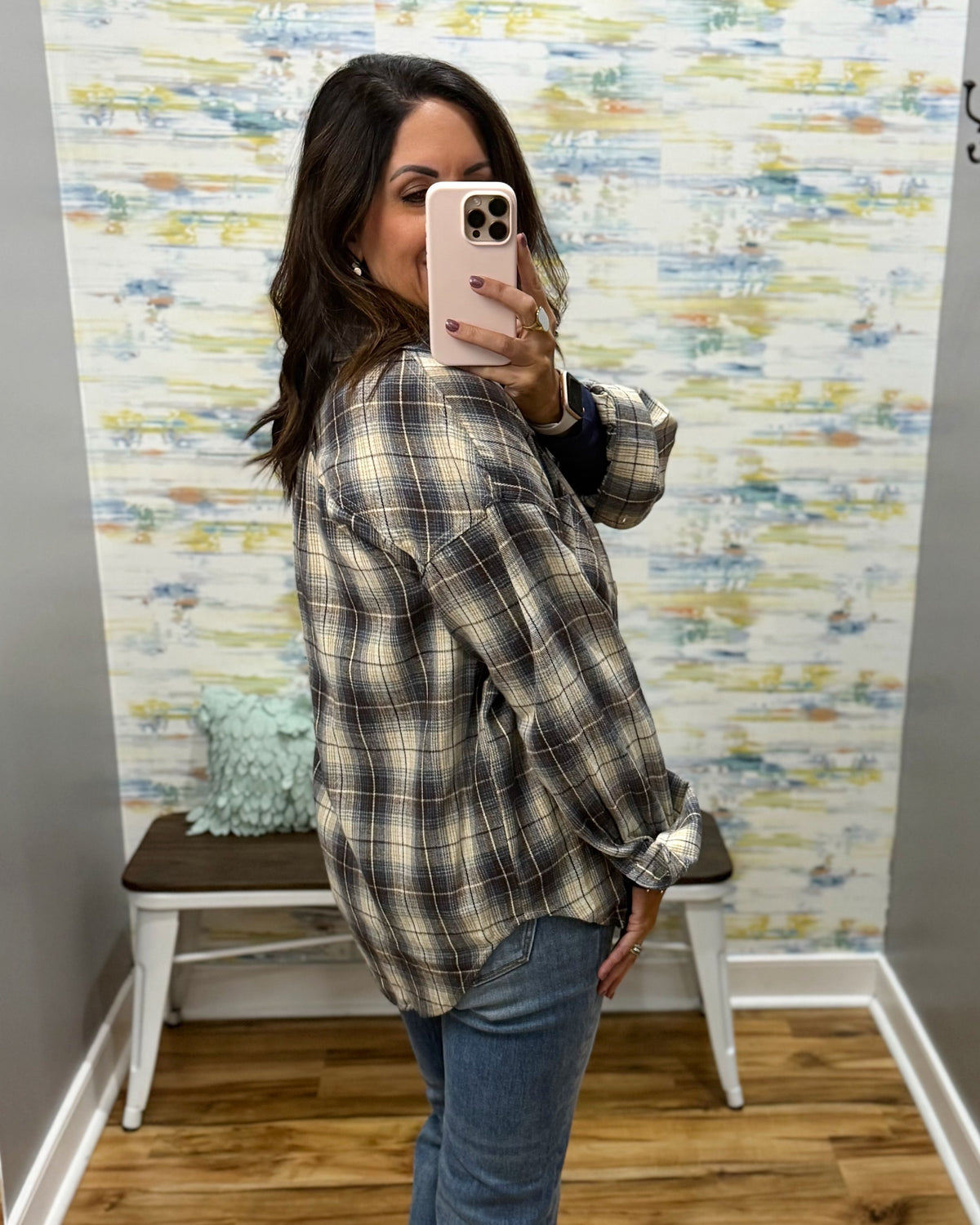 A woman takes a mirror selfie wearing an oversized plaid flannel shirt in beige, brown, and black, paired with blue jeans, in front of a decorative wall with a bench.