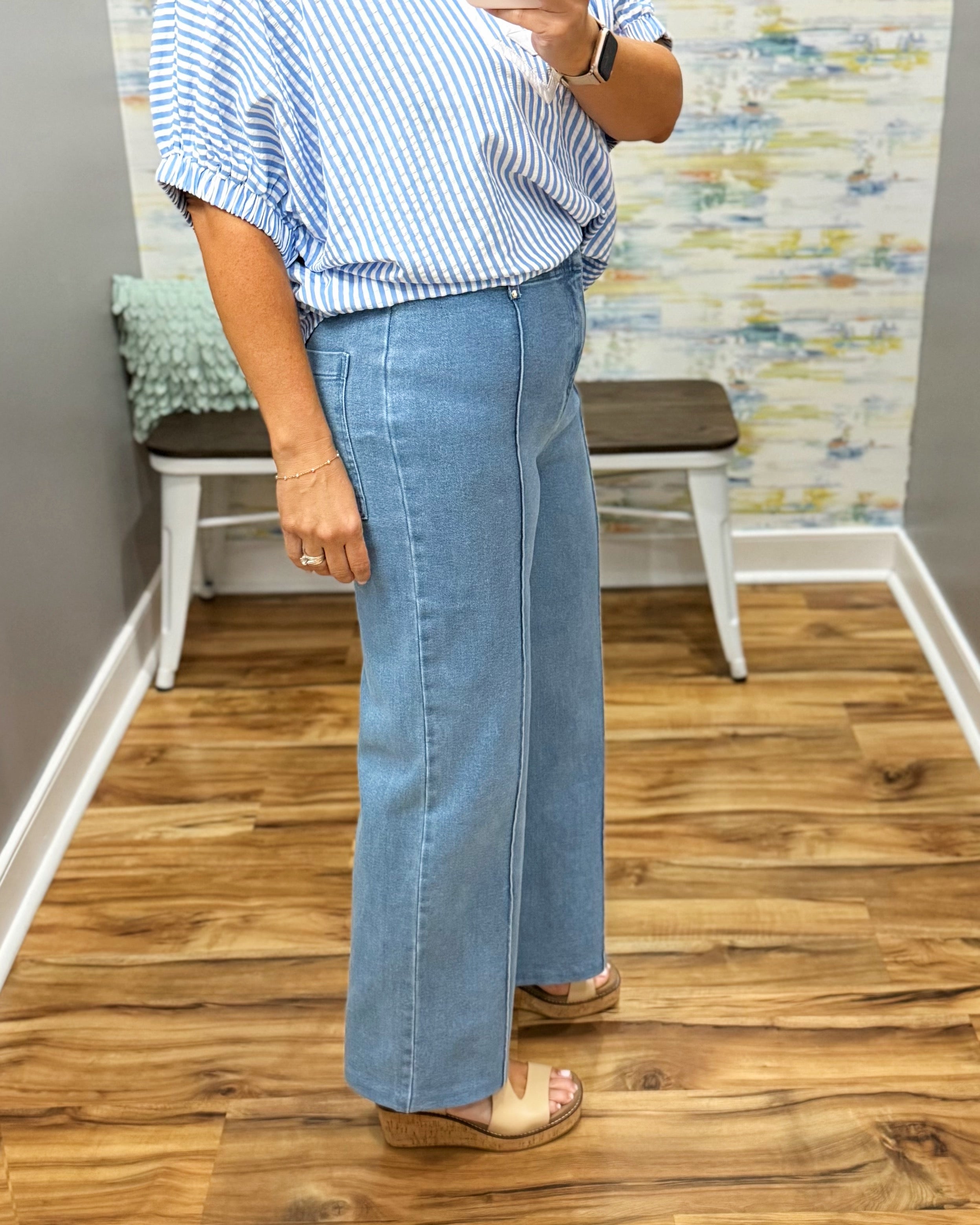 Person wearing a blue and white striped shirt and light blue jeans standing in a room with wooden flooring and a colorful wall.