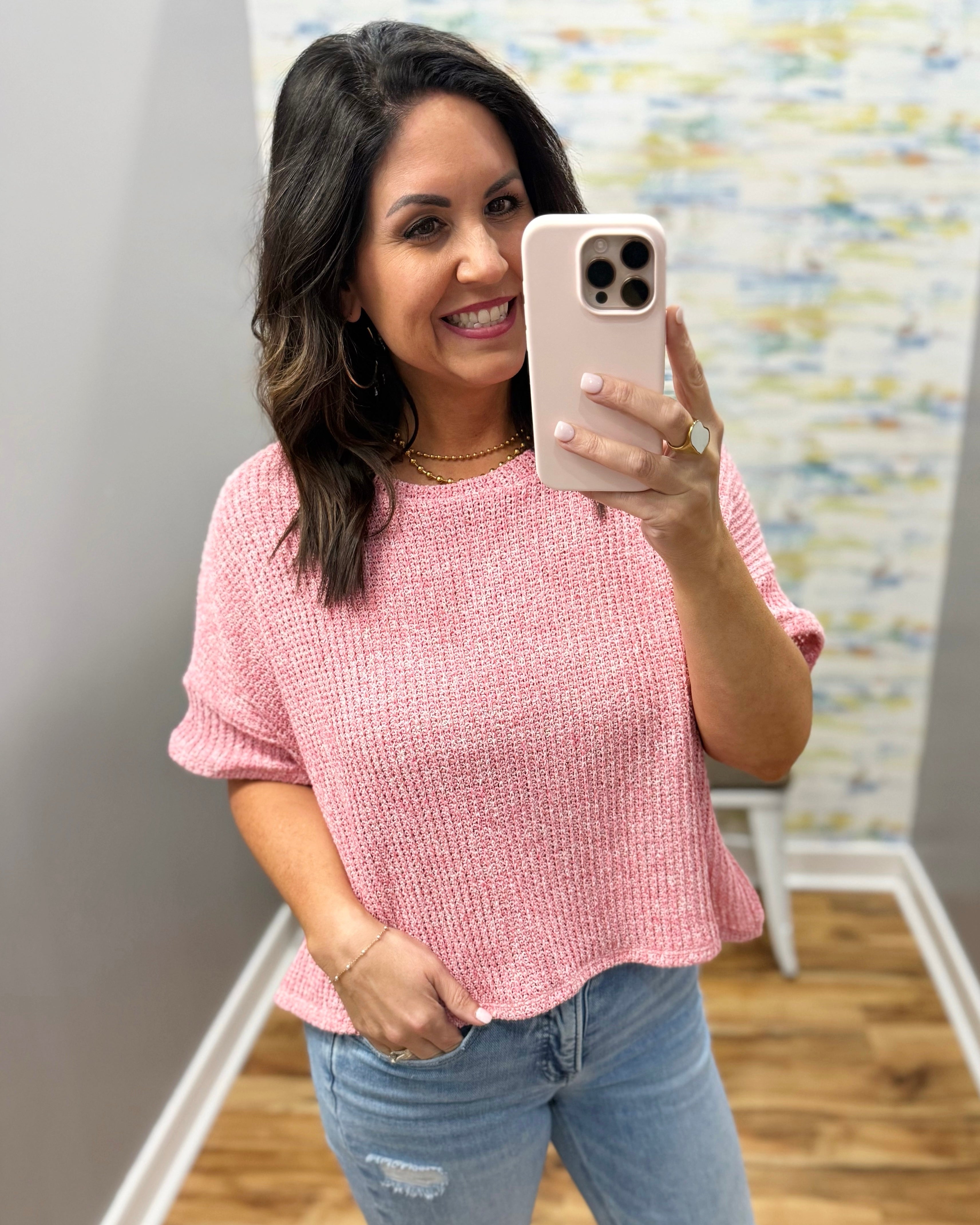 Woman taking a mirror selfie wearing a pink top and ripped jeans in a room with a colorful wall.